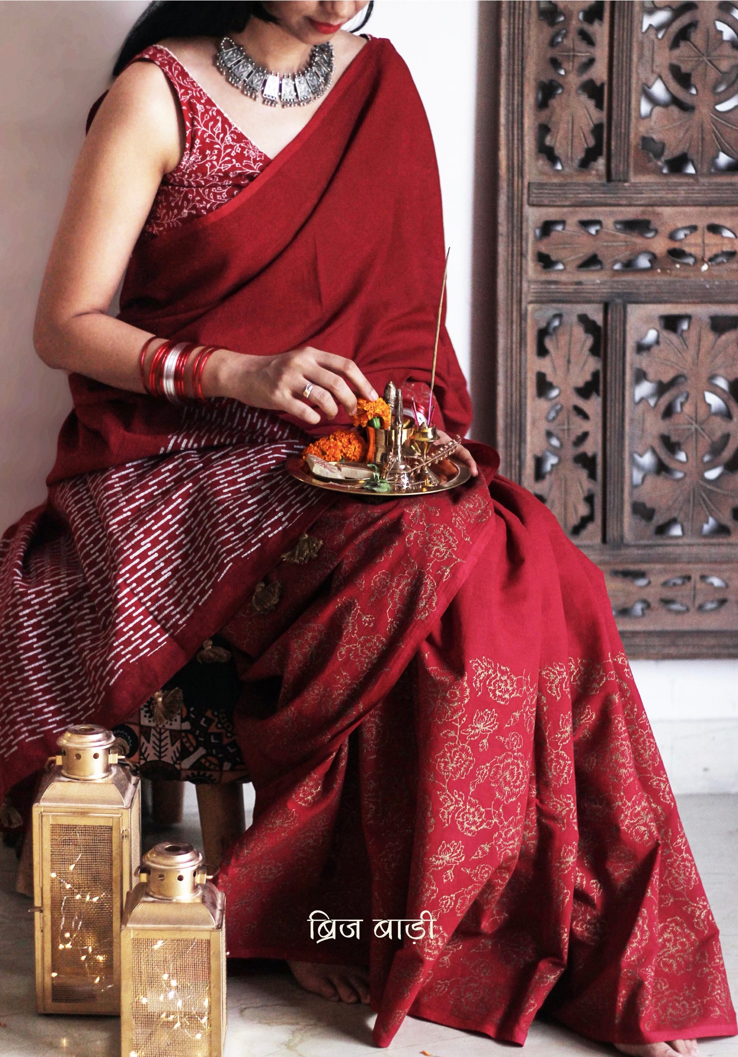 Deep red light-weight cotton saree by Brij Bari, featuring intricate rose block printed motifs in golden on the skirt and elegant geometric patterns on the pallu in silver, lending a festive and graceful touch. The pallu is finished with handcrafted olive green thread tassels that add earthy contrast and texture. Includes a free hand block printed blouse piece in matching tones. A perfect blend of festive elegance and artisan detail, crafted with care by Brij Bari.