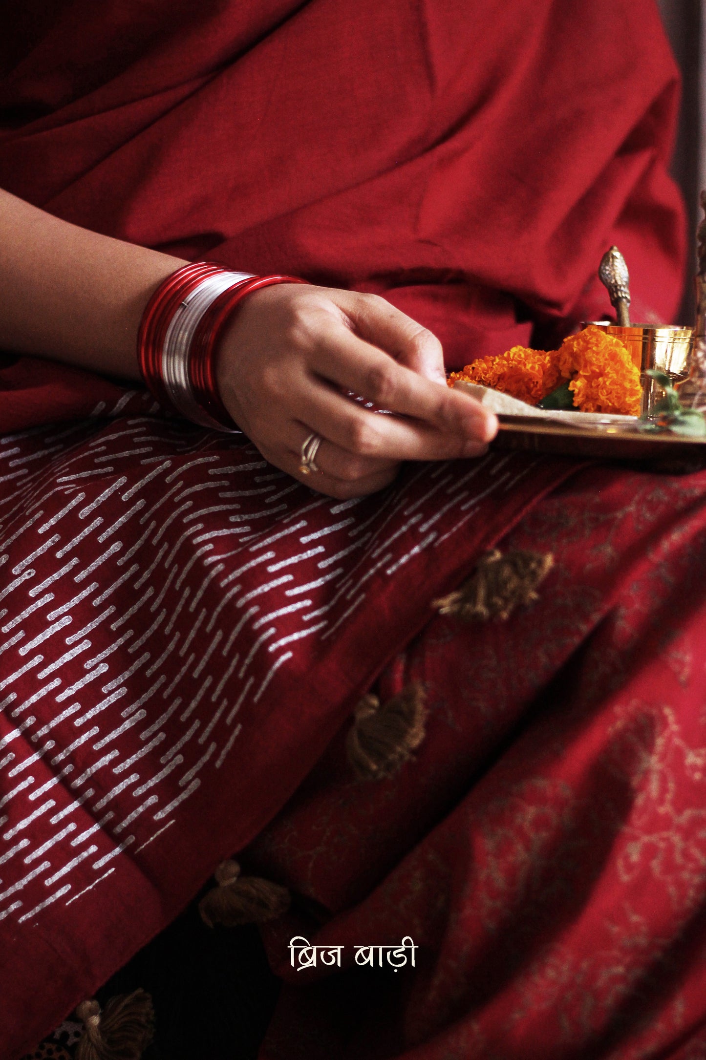 Deep red light-weight cotton saree by Brij Bari, featuring intricate rose block printed motifs in golden on the skirt and elegant geometric patterns on the pallu in silver, lending a festive and graceful touch. The pallu is finished with handcrafted olive green thread tassels that add earthy contrast and texture. Includes a free hand block printed blouse piece in matching tones. A perfect blend of festive elegance and artisan detail, crafted with care by Brij Bari.