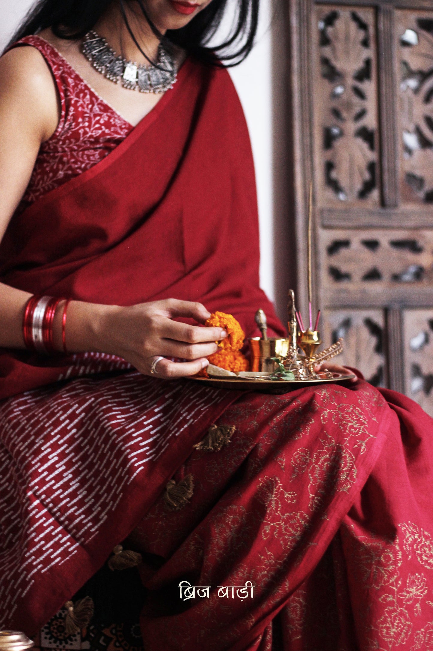 Deep red light-weight cotton saree by Brij Bari, featuring intricate rose block printed motifs in golden on the skirt and elegant geometric patterns on the pallu in silver, lending a festive and graceful touch. The pallu is finished with handcrafted olive green thread tassels that add earthy contrast and texture. Includes a free hand block printed blouse piece in matching tones. A perfect blend of festive elegance and artisan detail, crafted with care by Brij Bari.