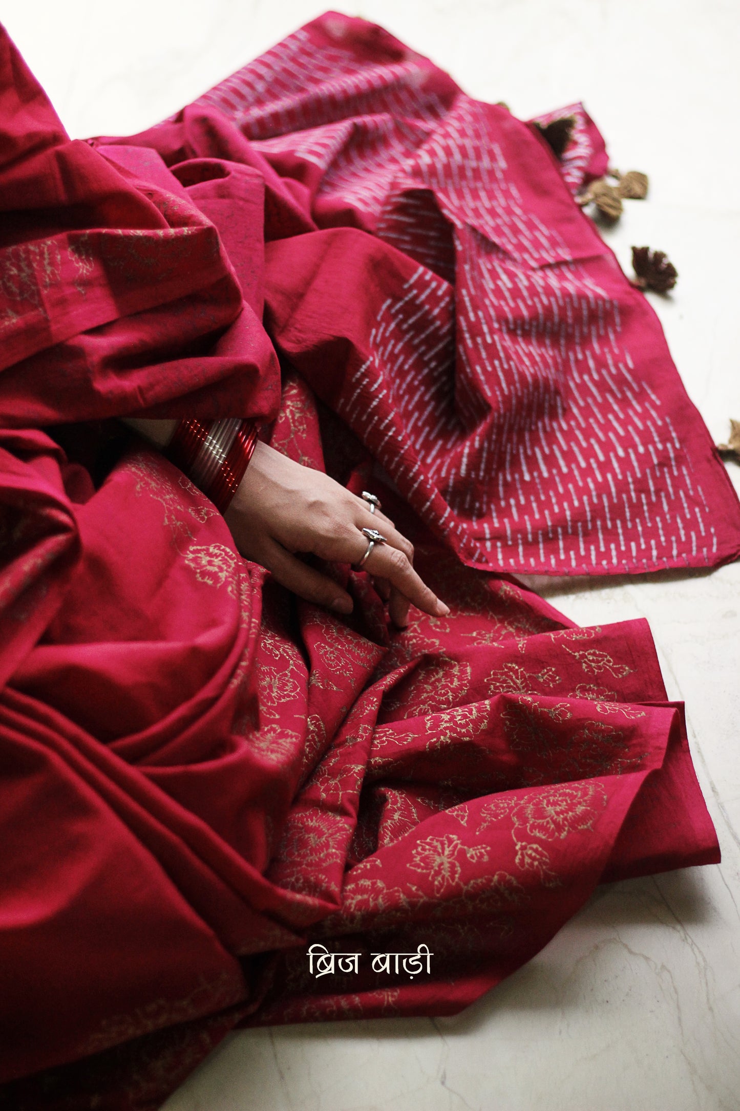 Deep red light-weight cotton saree by Brij Bari, featuring intricate rose block printed motifs in golden on the skirt and elegant geometric patterns on the pallu in silver, lending a festive and graceful touch. The pallu is finished with handcrafted olive green thread tassels that add earthy contrast and texture. Includes a free hand block printed blouse piece in matching tones. A perfect blend of festive elegance and artisan detail, crafted with care by Brij Bari.