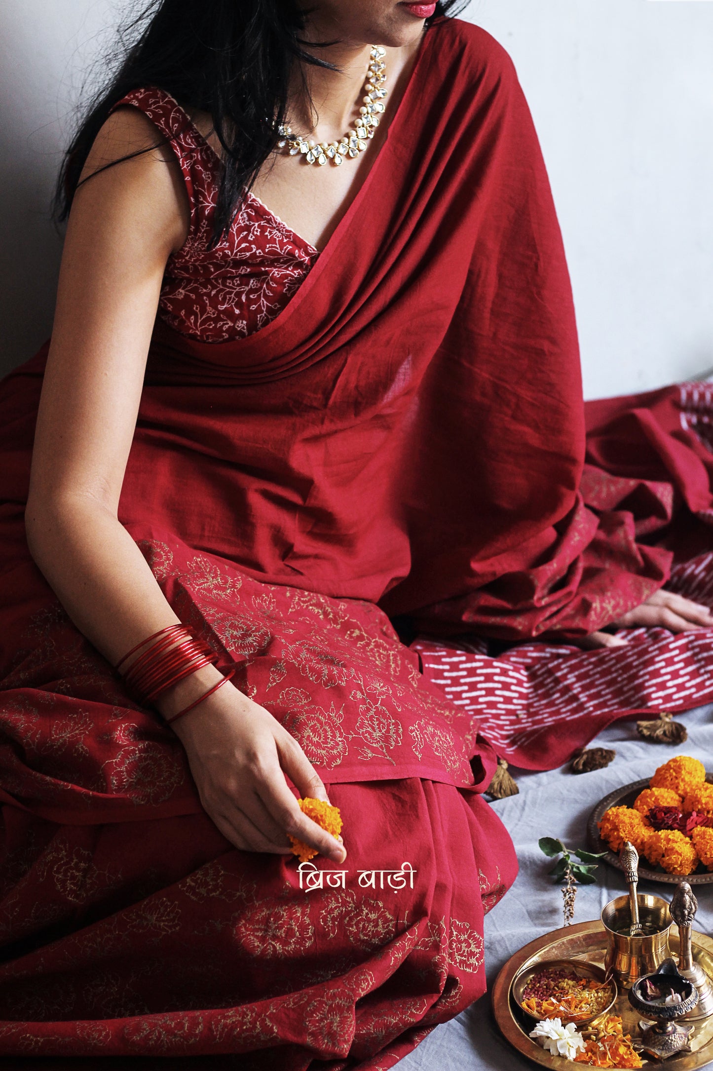 Deep red light-weight cotton saree by Brij Bari, featuring intricate rose block printed motifs in golden on the skirt and elegant geometric patterns on the pallu in silver, lending a festive and graceful touch. The pallu is finished with handcrafted olive green thread tassels that add earthy contrast and texture. Includes a free hand block printed blouse piece in matching tones. A perfect blend of festive elegance and artisan detail, crafted with care by Brij Bari.