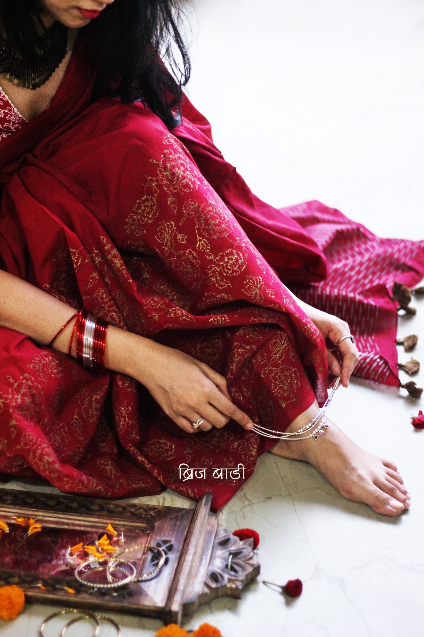 Deep red light-weight cotton saree by Brij Bari, featuring intricate rose block printed motifs in golden on the skirt and elegant geometric patterns on the pallu in silver, lending a festive and graceful touch. The pallu is finished with handcrafted olive green thread tassels that add earthy contrast and texture. Includes a free hand block printed blouse piece in matching tones. A perfect blend of festive elegance and artisan detail, crafted with care by Brij Bari.