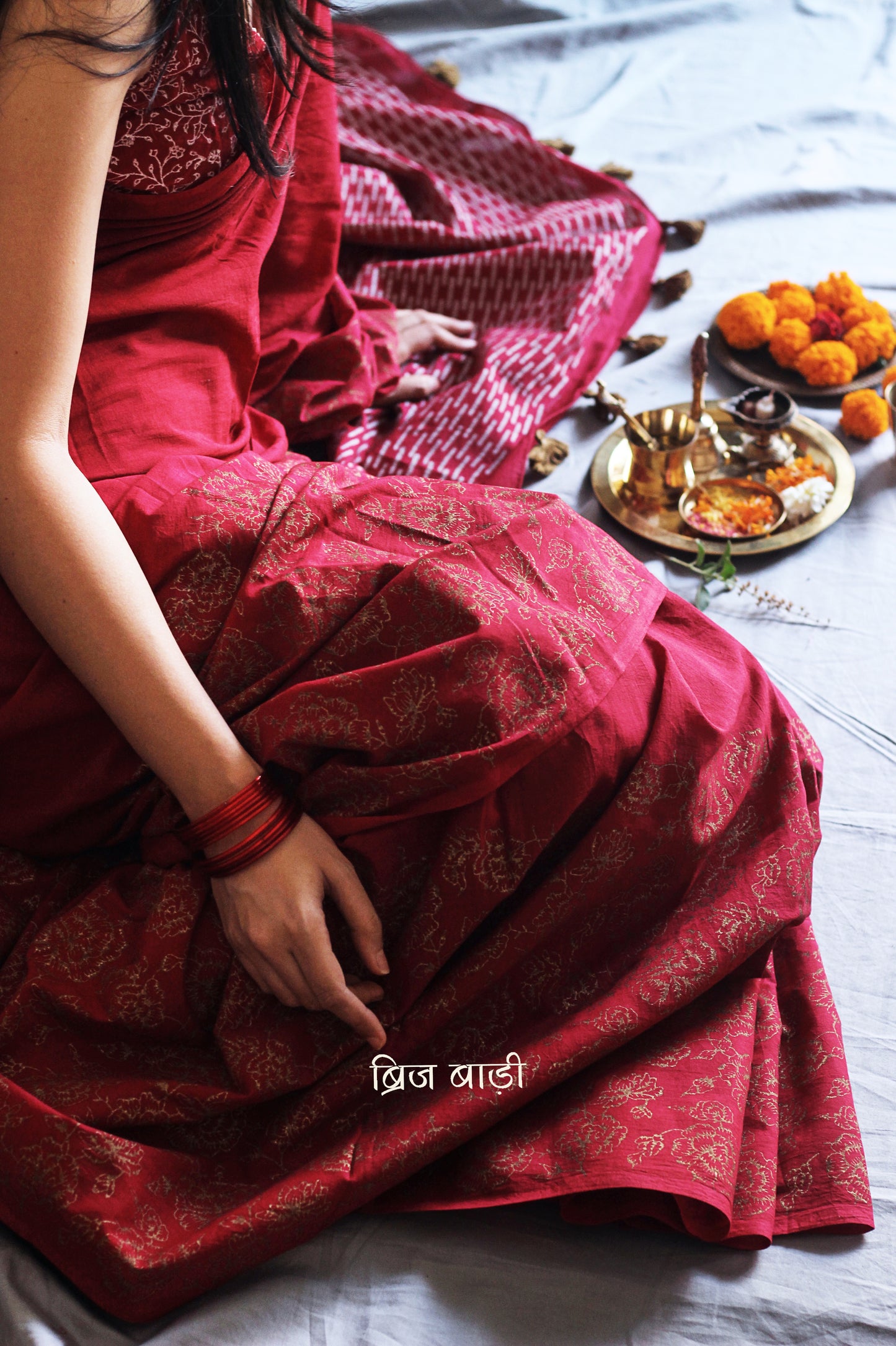 Deep red light-weight cotton saree by Brij Bari, featuring intricate rose block printed motifs in golden on the skirt and elegant geometric patterns on the pallu in silver, lending a festive and graceful touch. The pallu is finished with handcrafted olive green thread tassels that add earthy contrast and texture. Includes a free hand block printed blouse piece in matching tones. A perfect blend of festive elegance and artisan detail, crafted with care by Brij Bari.