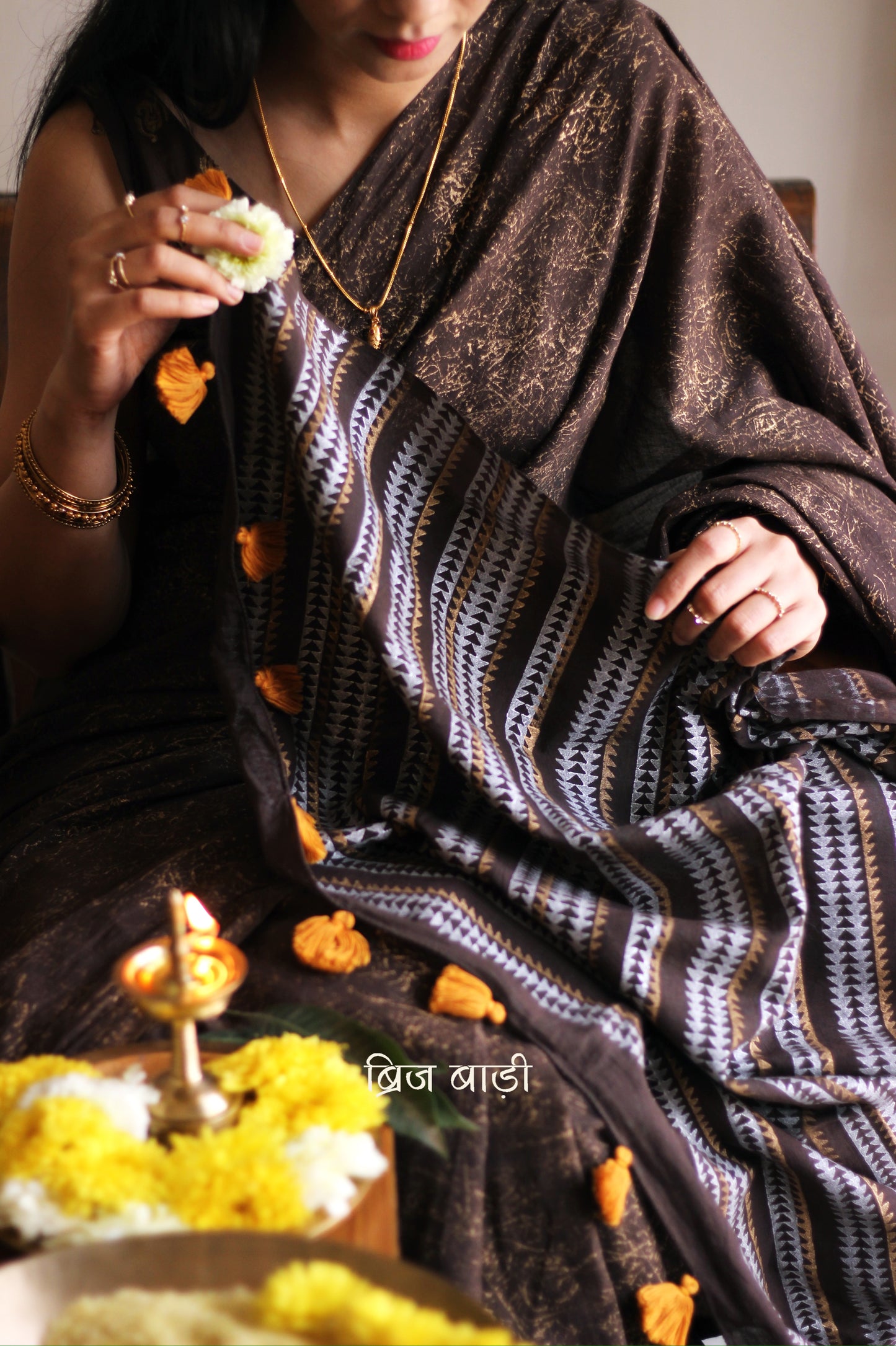Brown soft cotton saree by Brij Bari featuring a striking hand block printed abstract pattern in gold across the body, paired with a contemporary geometric design in silver and gold on the pallu. This blend of traditional technique and modern aesthetics gives the saree a unique festive appeal. The pallu is finished with handcrafted mustard thread tassels that add a warm, artisanal touch. Includes a free matching hand block printed blouse piece. A graceful and comfortable choice for festive gatherings