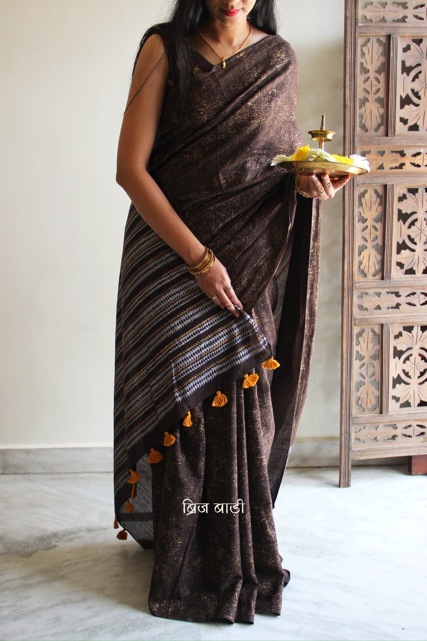Brown soft cotton saree by Brij Bari featuring a striking hand block printed abstract pattern in gold across the body, paired with a contemporary geometric design in silver and gold on the pallu. This blend of traditional technique and modern aesthetics gives the saree a unique festive appeal. The pallu is finished with handcrafted mustard thread tassels that add a warm, artisanal touch. Includes a free matching hand block printed blouse piece. A graceful and comfortable choice for festive gatherings