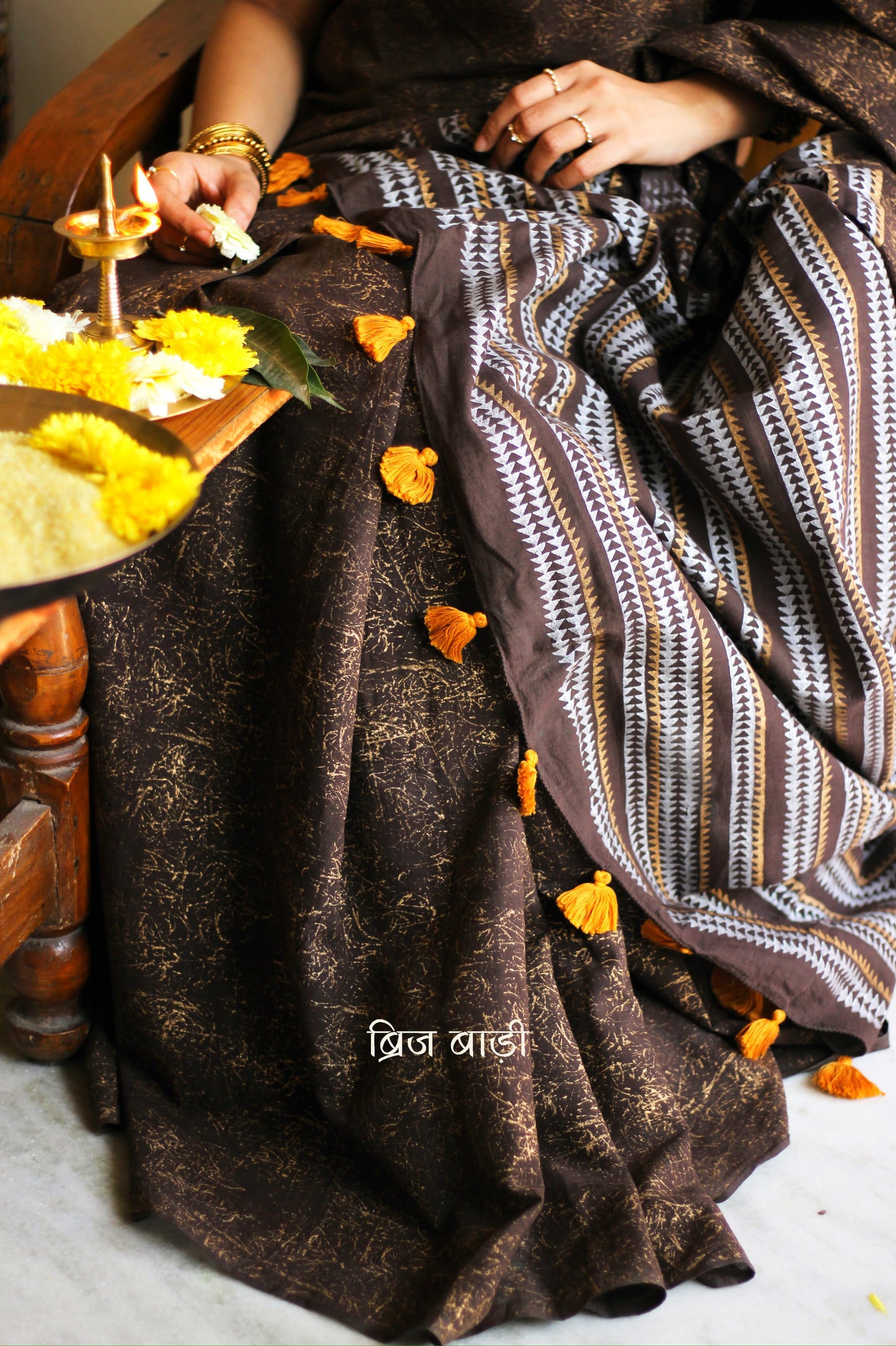 Brown soft cotton saree by Brij Bari featuring a striking hand block printed abstract pattern in gold across the body, paired with a contemporary geometric design in silver and gold on the pallu. This blend of traditional technique and modern aesthetics gives the saree a unique festive appeal. The pallu is finished with handcrafted mustard thread tassels that add a warm, artisanal touch. Includes a free matching hand block printed blouse piece. A graceful and comfortable choice for festive gatherings