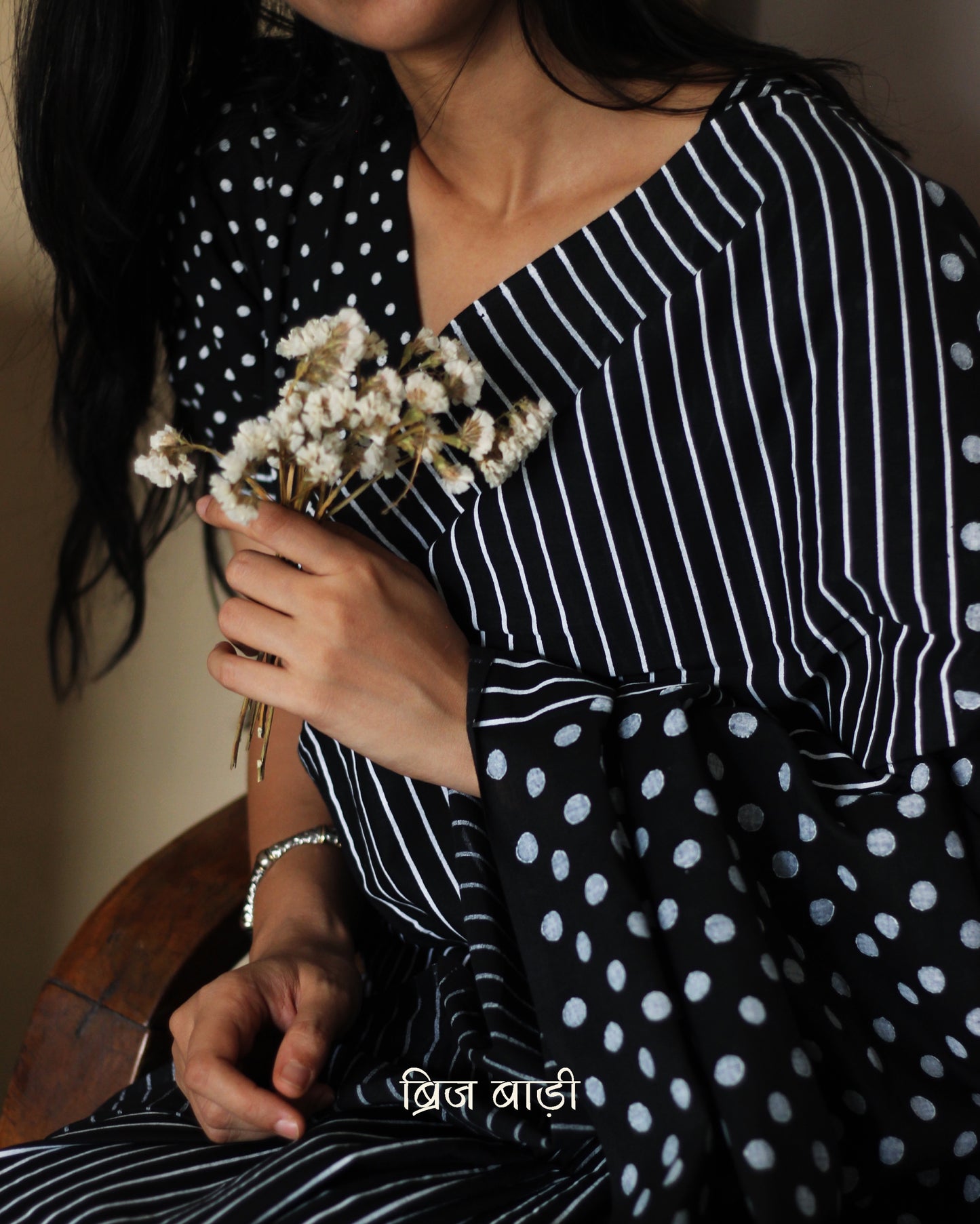 Black soft cotton saree by Brij Bari, featuring a bold and striking hand block printed pattern of stripes and polka dots for a modern, playful twist. Crafted from breathable, light-weight cotton, the saree drapes effortlessly—ideal for both everyday wear and special occasions. Finished with handcrafted red thread tassels on the pallu, adding an artisanal touch of charm. It comes with a free hand block printed blouse piece that complements the print and palette.