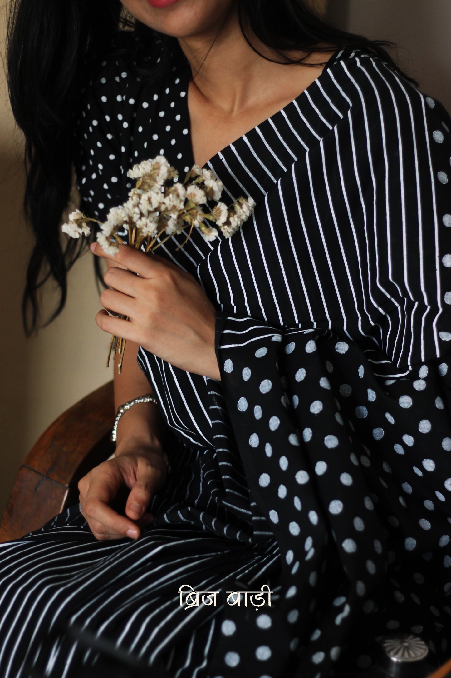 Black soft cotton saree by Brij Bari, featuring a bold and striking hand block printed pattern of stripes and polka dots for a modern, playful twist. Handcrafted from breathable, light-weight cotton, the saree drapes effortlessly—ideal for both everyday wear and special occasions. Finished with handcrafted red thread tassels on the pallu, adding an artisanal touch of charm. It comes with a free hand block printed blouse piece that complements the print and palette.