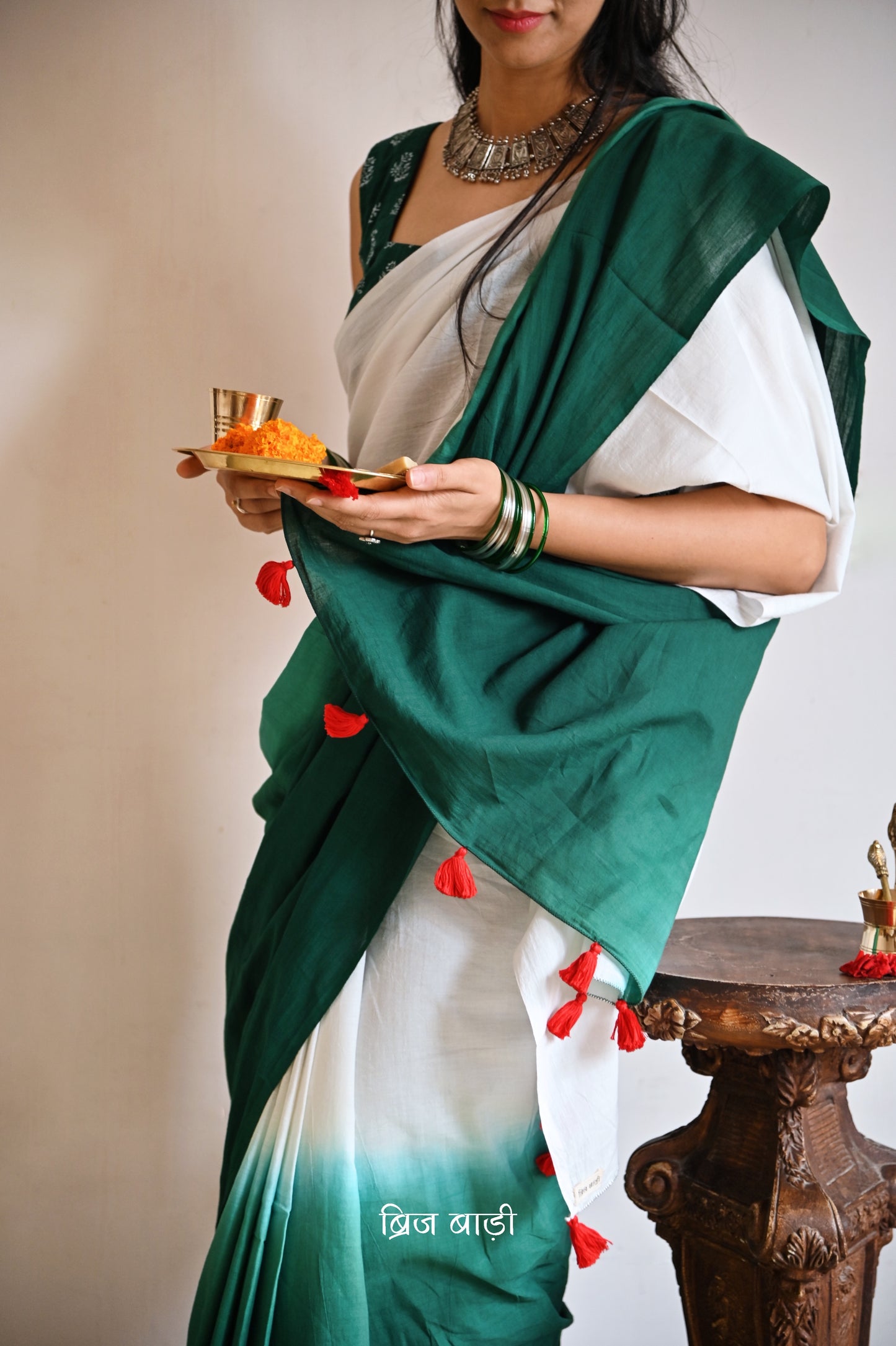 Dark Green and off-white ombré soft cotton saree by Brij Bari, crafted for effortless elegance and all-day comfort. The subtle blend of tones adds fluidity and grace to the drape, making it perfect for both everyday wear and special occasions. The pallu is adorned with handcrafted red thread tassels, lending a warm, artisanal touch. It comes with a free hand block printed blouse piece that complements the saree’s minimal yet striking aesthetic. Available in 8 beautiful colour options