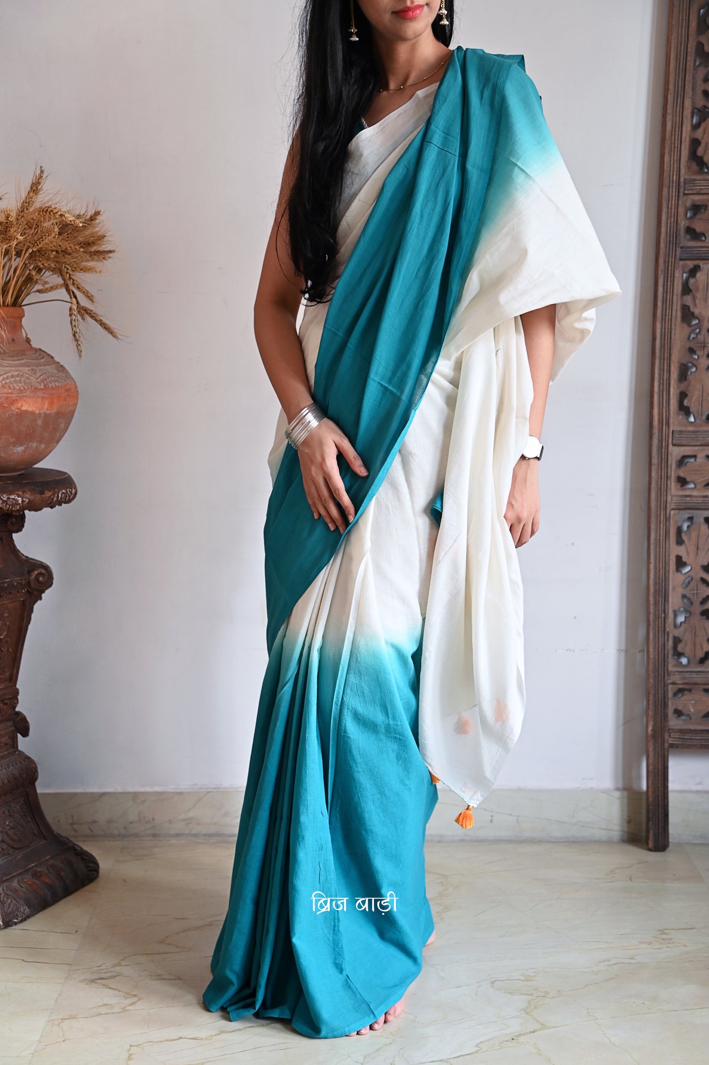 Teal Blue and off-white ombré soft cotton saree by Brij Bari, crafted for effortless elegance and all-day comfort. The subtle blend of tones adds fluidity and grace to the drape, making it perfect for both everyday wear and special occasions. The pallu is adorned with handcrafted orange thread tassels, lending a warm, artisanal touch. It comes with a free hand block printed blouse piece that complements the saree’s minimal yet striking aesthetic. Available in 8 beautiful colour options