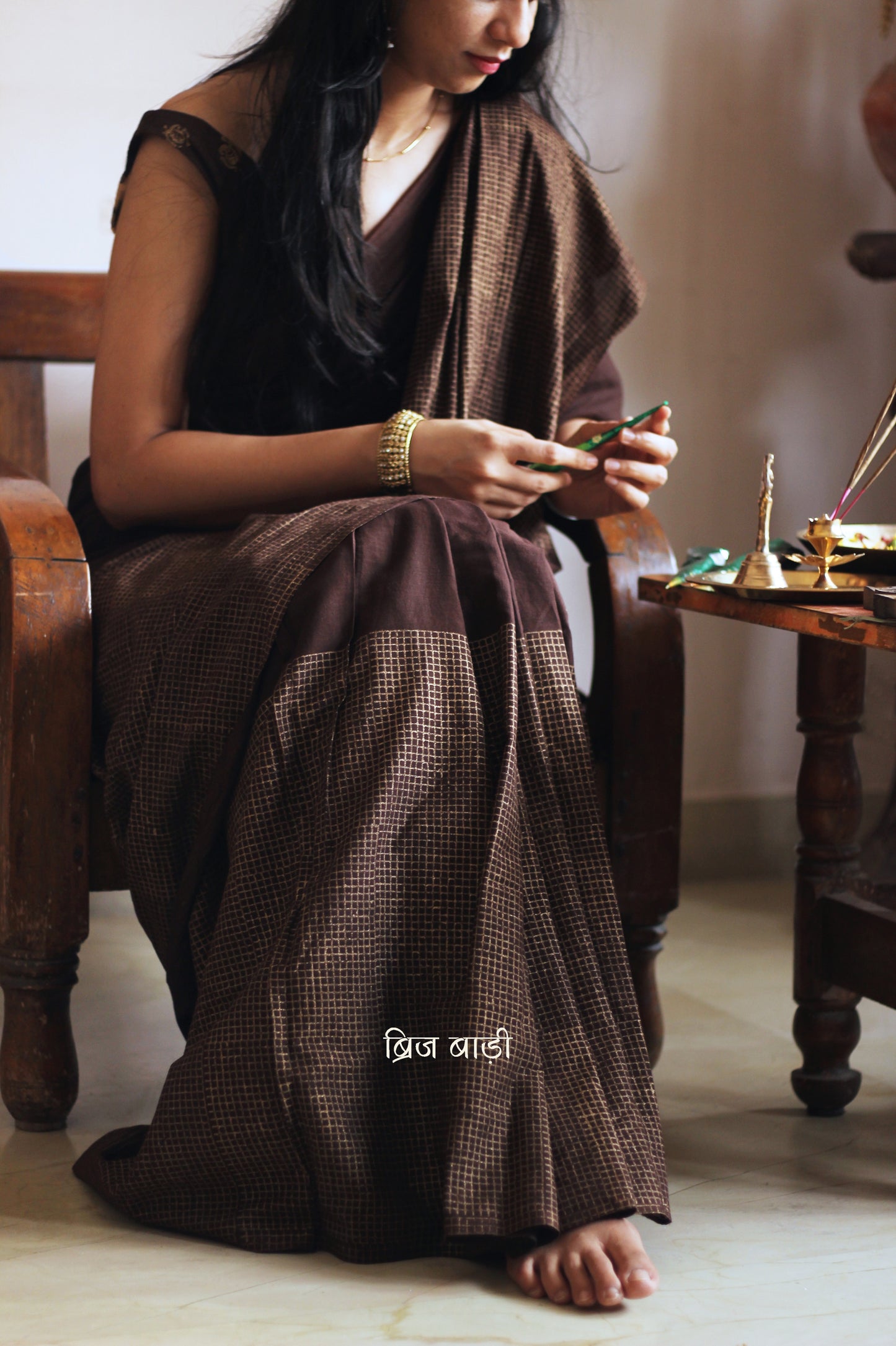 Earthy brown light-weight cotton saree by Brij Bari, featuring intricate golden hand block printed check patterns on the skirt and silver geometric designs on the pallu. Handcrafted mustard thread tassels add a warm, artisanal touch to the pallu. Includes a free hand block printed blouse piece that complements the saree’s festive and versatile appeal.