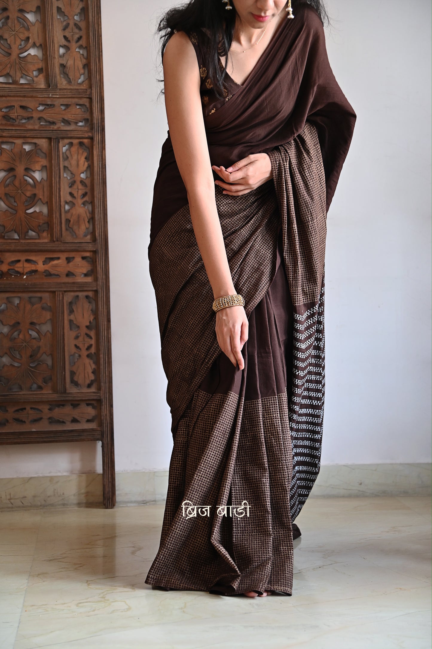 Earthy brown light-weight cotton saree by Brij Bari, featuring intricate golden hand block printed check patterns on the skirt and silver geometric designs on the pallu. Handcrafted mustard thread tassels add a warm, artisanal touch to the pallu. Includes a free hand block printed blouse piece that complements the saree’s festive and versatile appeal.