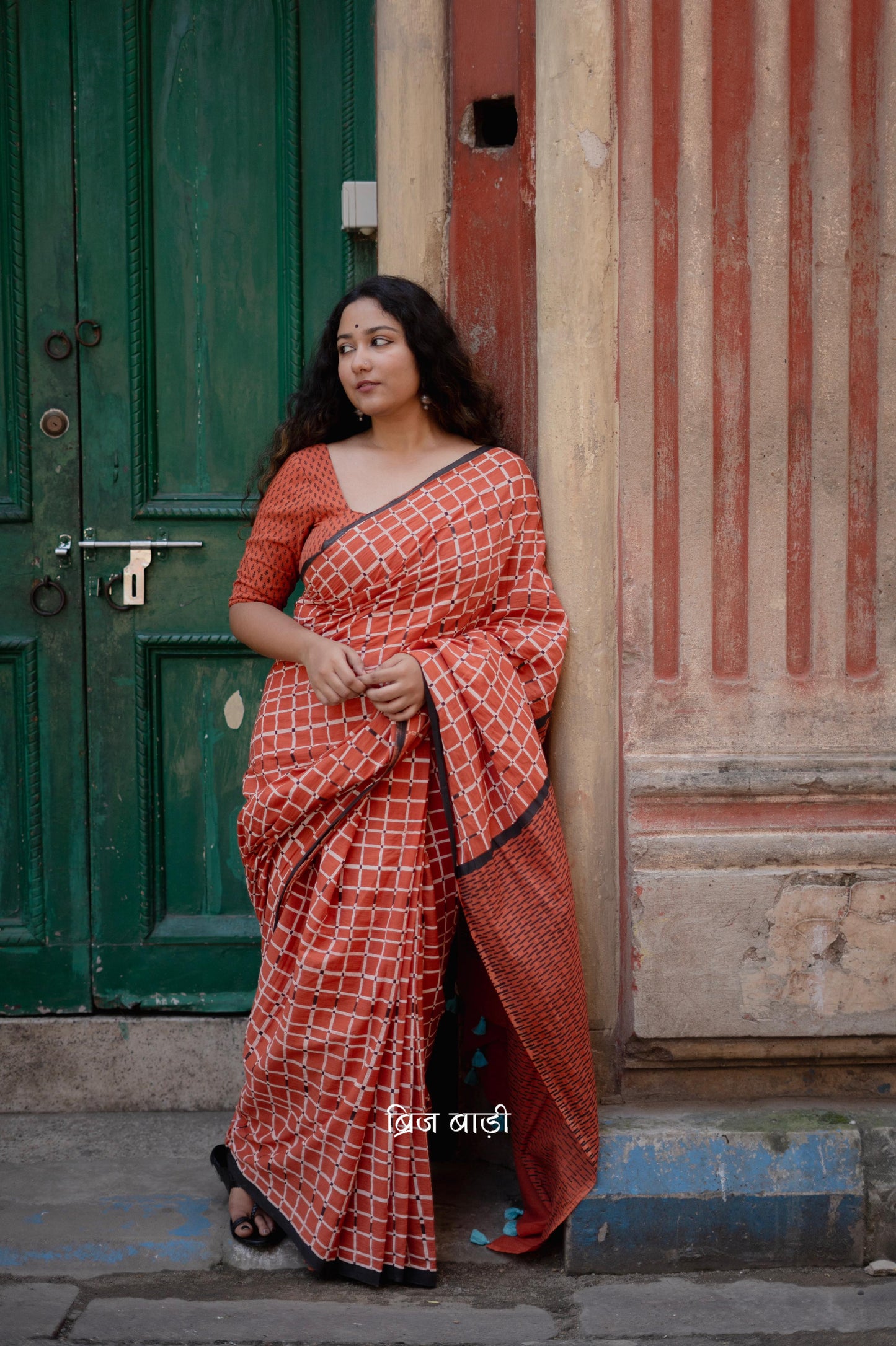 Earthy rust cotton saree by Brij Bari, hand block printed with black and white check pattern. Designed in soft, lightweight cotton with handmade light blue thread tassels on the pallu. Includes a free hand block printed cotton blouse piece. Perfect for adding a handcrafted, artisanal touch to your daily wear with breathable comfort and effortless style. Free shipping pan India.