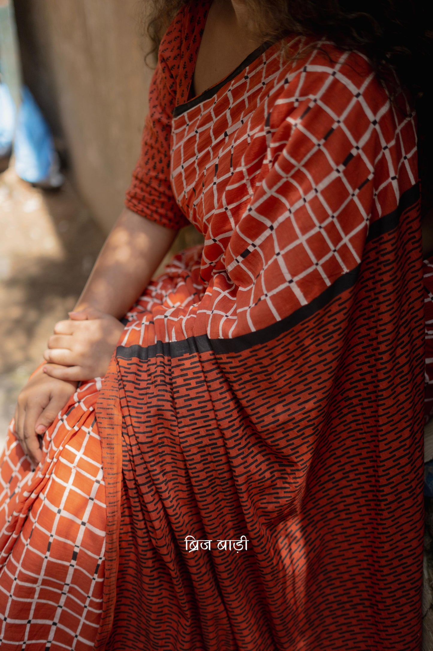 Earthy rust cotton saree by Brij Bari, hand block printed with black and white check pattern. Designed in soft, lightweight cotton with handmade light blue thread tassels on the pallu. Includes a free hand block printed cotton blouse piece. Perfect for adding a handcrafted, artisanal touch to your daily wear with breathable comfort and effortless style. Free shipping pan India.