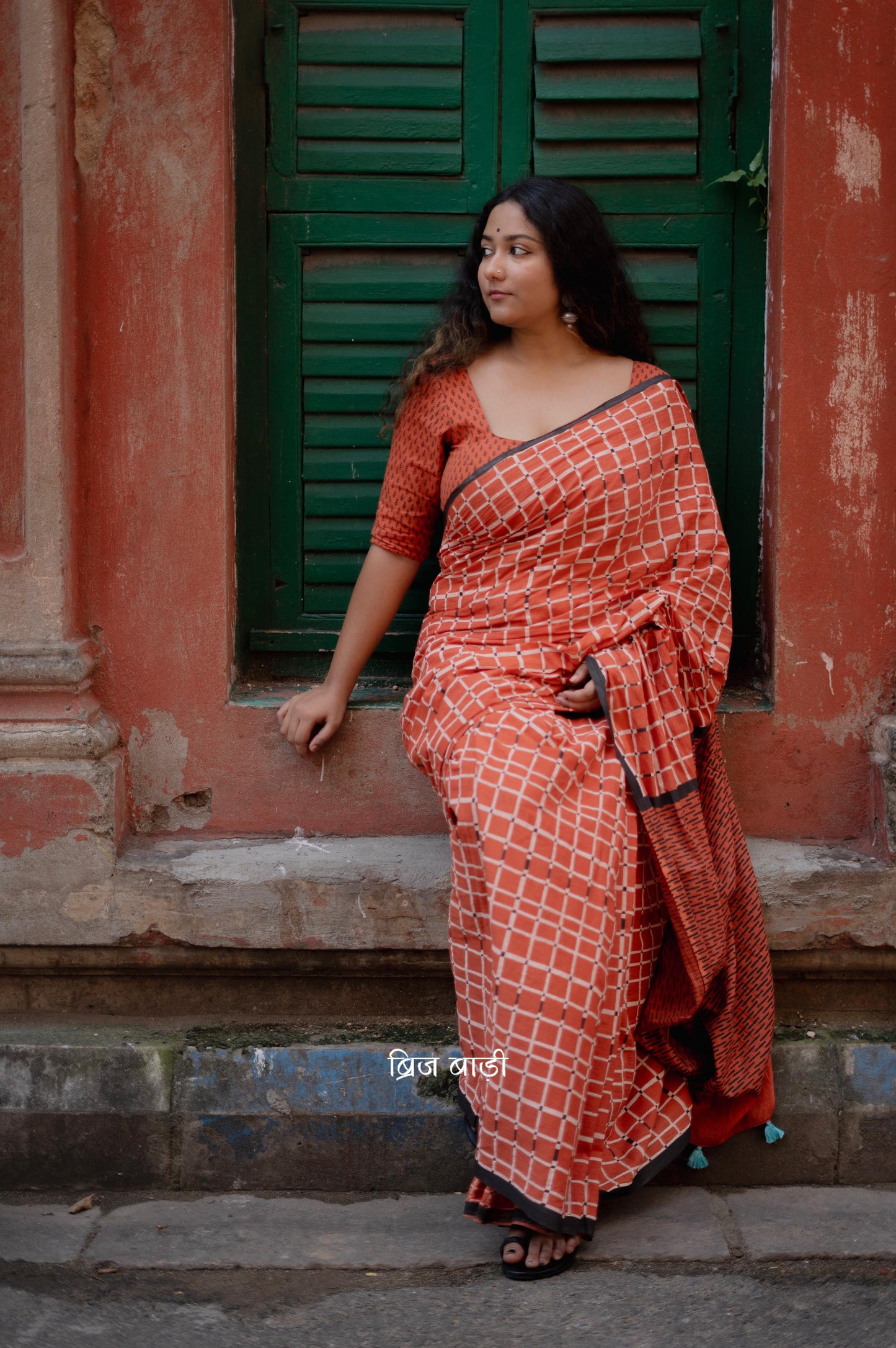 Earthy rust cotton saree by Brij Bari, hand block printed with black and white check pattern. Designed in soft, lightweight cotton with handmade light blue thread tassels on the pallu. Includes a free hand block printed cotton blouse piece. Perfect for adding a handcrafted, artisanal touch to your daily wear with breathable comfort and effortless style. Free shipping pan India.