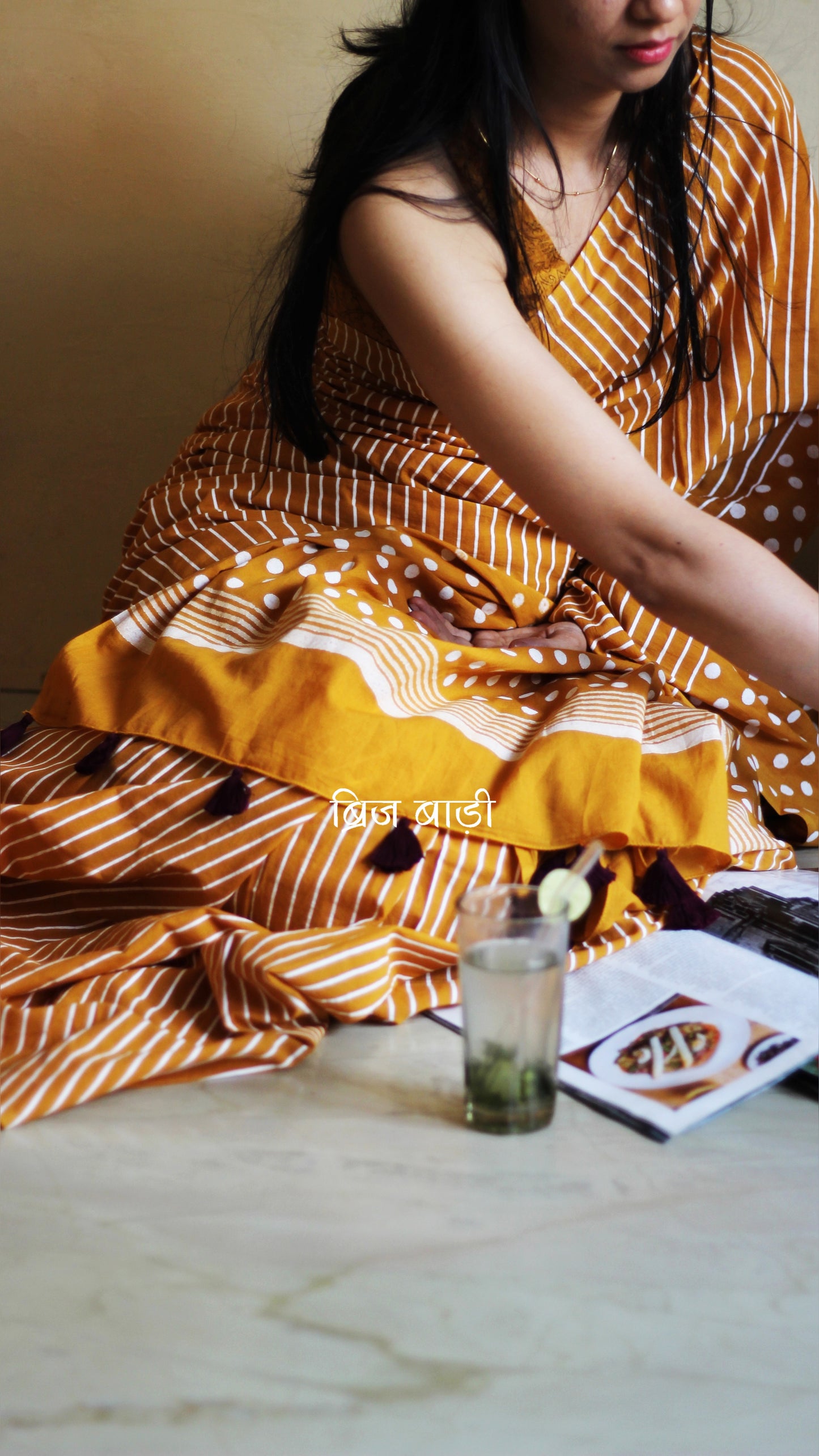 Yellow soft cotton saree by Brij Bari, featuring a bold and striking hand block printed pattern of stripes and polka dots for a modern, playful twist. Crafted from breathable, light-weight cotton, the saree drapes effortlessly—ideal for both everyday wear and special occasions. Finished with handcrafted purple thread tassels on the pallu, adding an artisanal touch of charm. It comes with a free hand block printed blouse piece that complements the print and palette.
