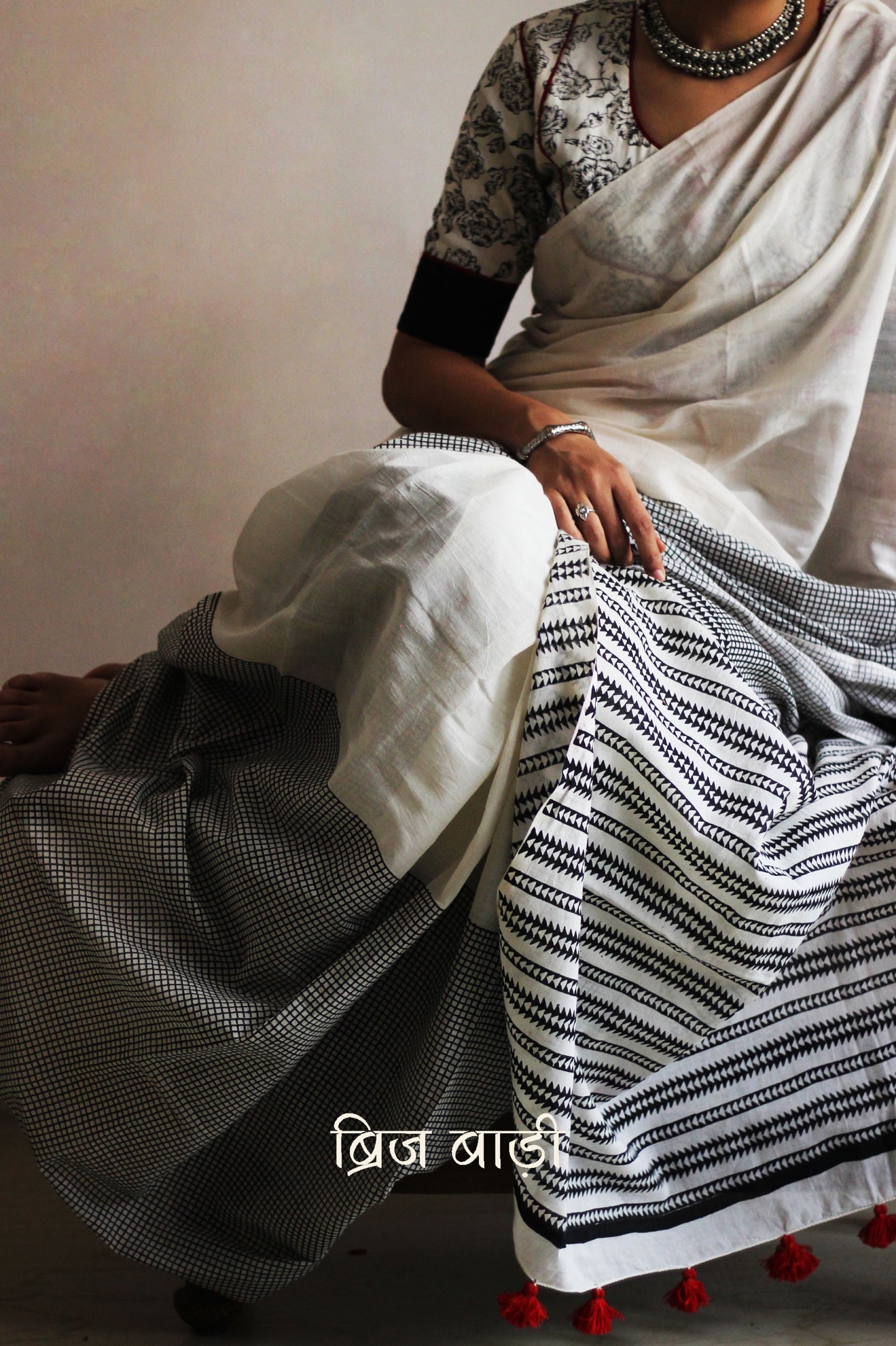 Off-white soft and light-weight cotton saree by Brij Bari, featuring a hand block printed check pattern on the skirt and geometrical pattern on the pallu. Designed for effortless everyday wear, this breathable, easy-drape saree brings together traditional craft with contemporary patterns. Finished with bright red handmade thread tassels on the pallu. Paired with a free hand block printed blouse piece that compliments the design perfectly. Offers free shipping pan India