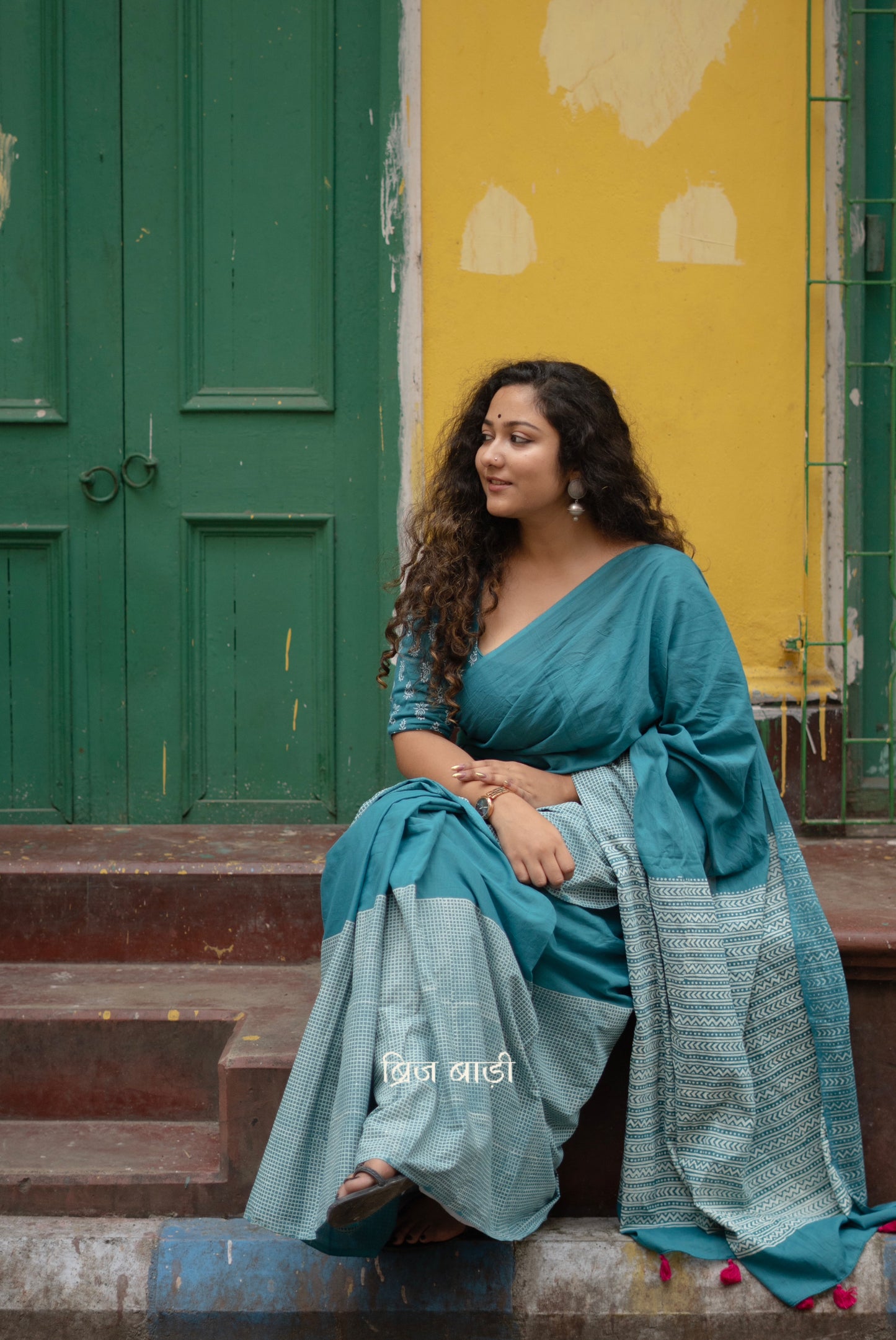 Teal blue soft and lightweight cotton saree by Brij Bari, hand block printed with a classic check pattern on the skirt and a geometric design in white on the pallu. Finished with handmade orange thread tassels on the pallu. Comes with a complimentary block printed cotton blouse piece. A versatile design that adds a pop of colour to your everyday wardrobe while offering breathable comfort and effortless style. Free shipping pan India.