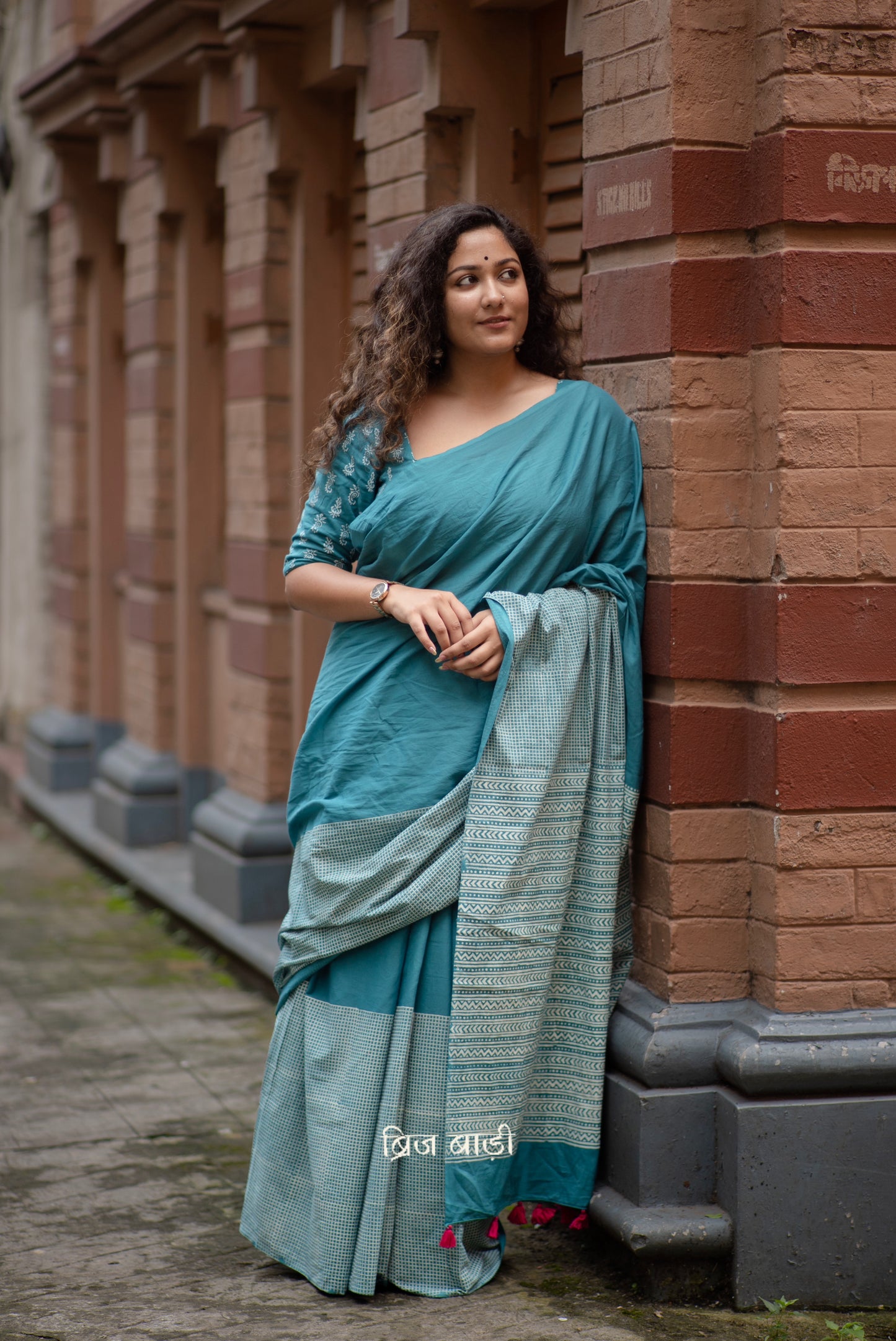 Teal blue soft and lightweight cotton saree by Brij Bari, hand block printed with a classic check pattern on the skirt and a geometric design in white on the pallu. Finished with handmade orange thread tassels on the pallu. Comes with a complimentary block printed cotton blouse piece. A versatile design that adds a pop of colour to your everyday wardrobe while offering breathable comfort and effortless style. Free shipping pan India.