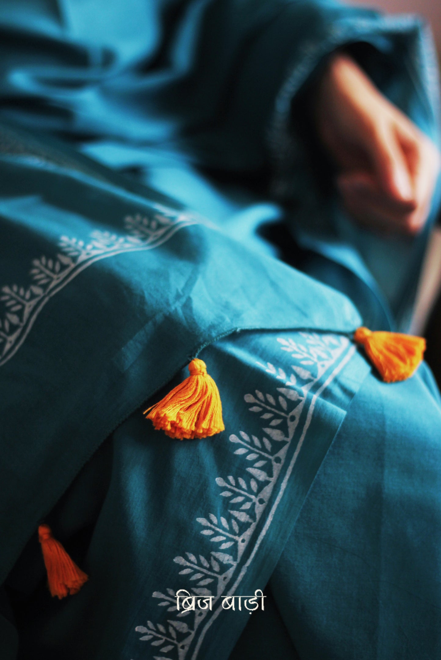 Teal blue soft and light-weight cotton saree by Brij Bari, designed for all-day comfort and effortless elegance. Detailed with a delicate hand block printed border in crisp white, creating a refined contrast. The saree is finished with orange handmade thread tassels on the pallu, adding a subtle pop of colour. Comes with a free matching block printed blouse piece. Ideal for everyday wear, office, casual outings, or light festive occasions, with free shipping pan India.