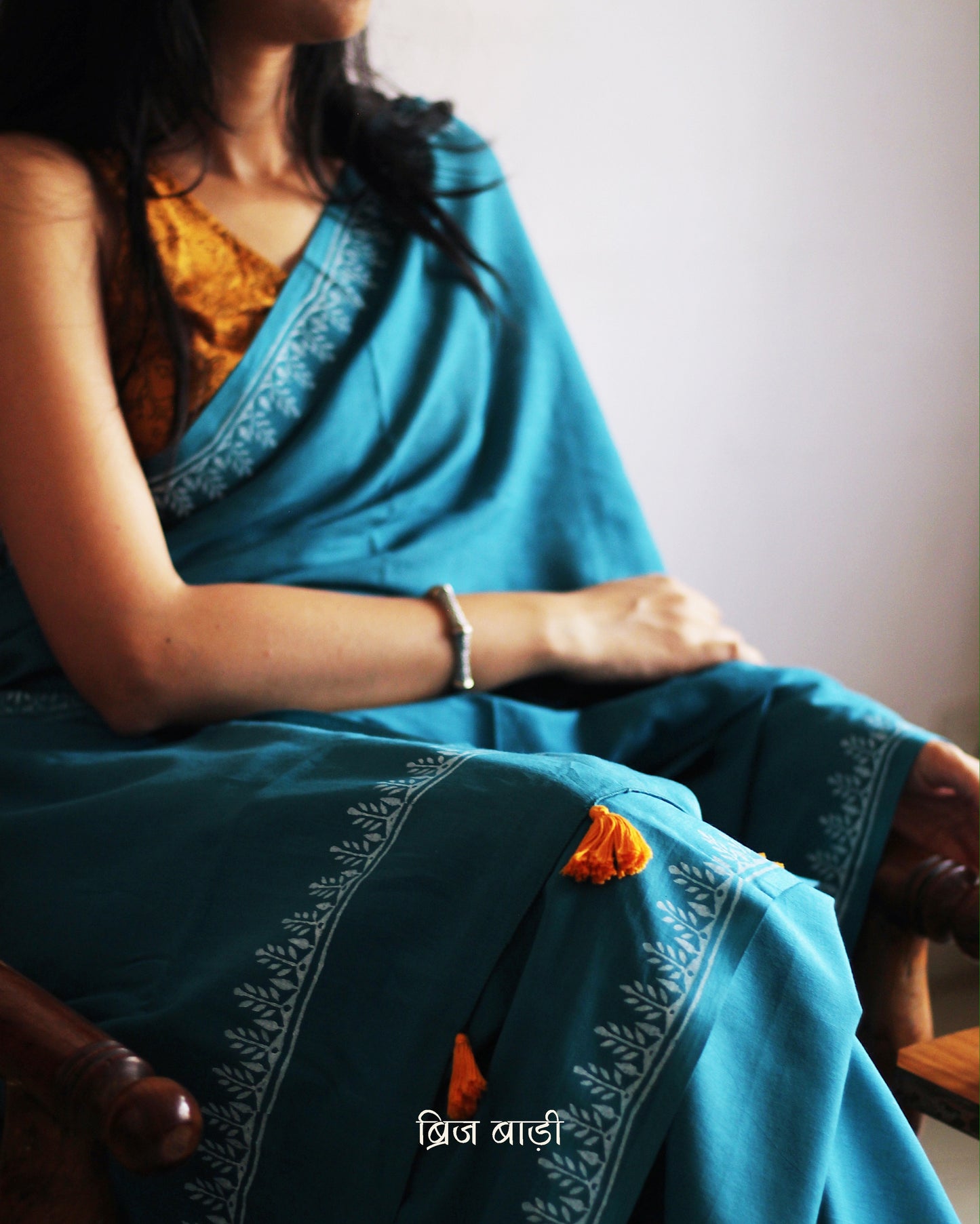 Teal blue soft and light-weight cotton saree by Brij Bari, designed for all-day comfort and effortless elegance. Detailed with a delicate hand block printed border in crisp white, creating a refined contrast. The saree is finished with orange handmade thread tassels on the pallu, adding a subtle pop of colour. Comes with a free matching block printed blouse piece. Ideal for everyday wear, office, casual outings, or light festive occasions, with free shipping pan India.