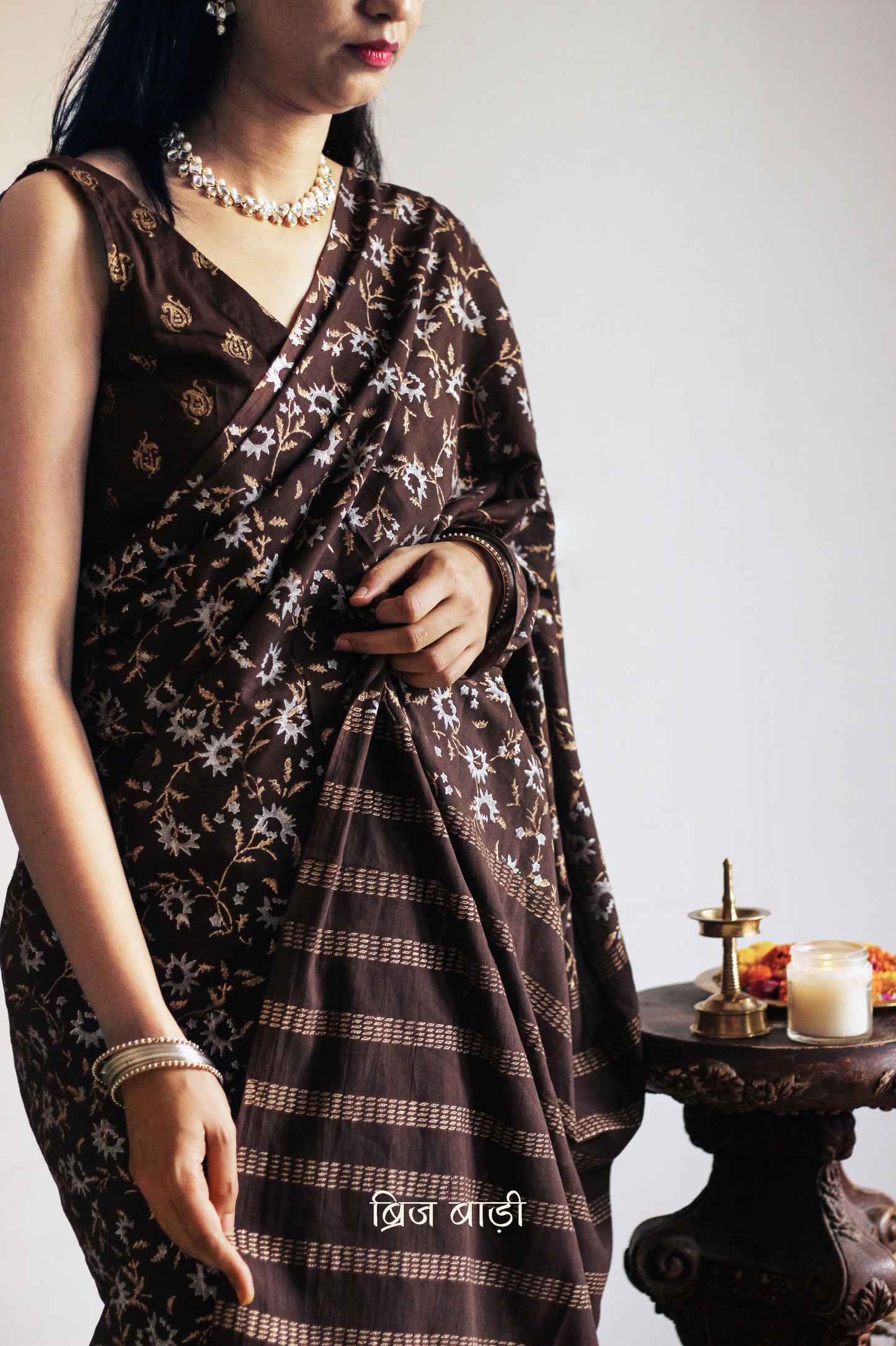 Brown soft cotton saree by Brij Bari, adorned with an abstract floral hand block print in elegant shades of gold and silver, lending it a refined festive charm. Crafted from breathable, light-weight cotton, the saree is accented with handcrafted mustard thread tassels on the pallu for a warm, artisanal finish. It comes with a free hand block printed blouse piece that complements the overall design. Ideal for festive occasions, this saree blends traditional techniques with modern aesthetics