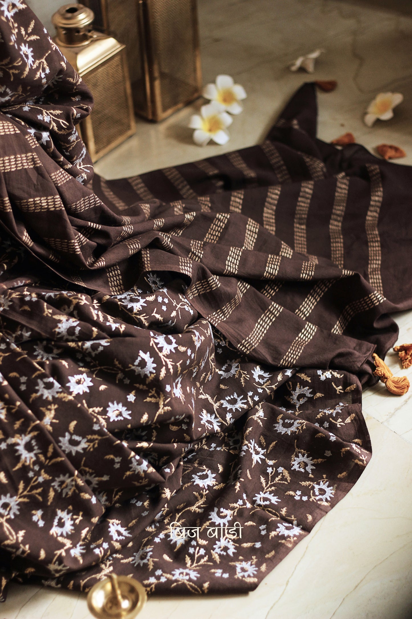 Brown soft cotton saree by Brij Bari, adorned with an abstract floral hand block print in elegant shades of gold and silver, lending it a refined festive charm. Crafted from breathable, light-weight cotton, the saree is accented with handcrafted mustard thread tassels on the pallu for a warm, artisanal finish. It comes with a free hand block printed blouse piece that complements the overall design. Ideal for festive occasions, this saree blends traditional techniques with modern aesthetics