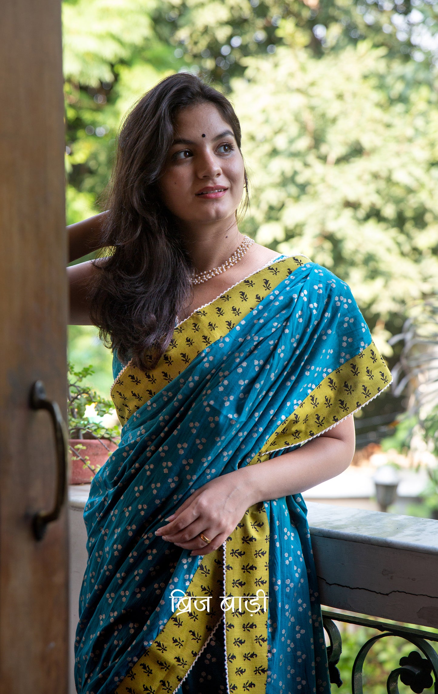 Teal blue cotton saree by Brij Bari, hand block printed with silver polka dot motifs & detailed with contrasting light green block printed border. Accented with delicate crochet lace that adds to its handcrafted charm. The pallu is finished with artisanal yellow & pink handmade thread tassels & tiny sea shells on the corner infusing it with a joyful festive flair. Comes with a free block printed blouse piece. A perfect blend of comfort & striking elegance for festive gatherings. Free blouse piece & shipping