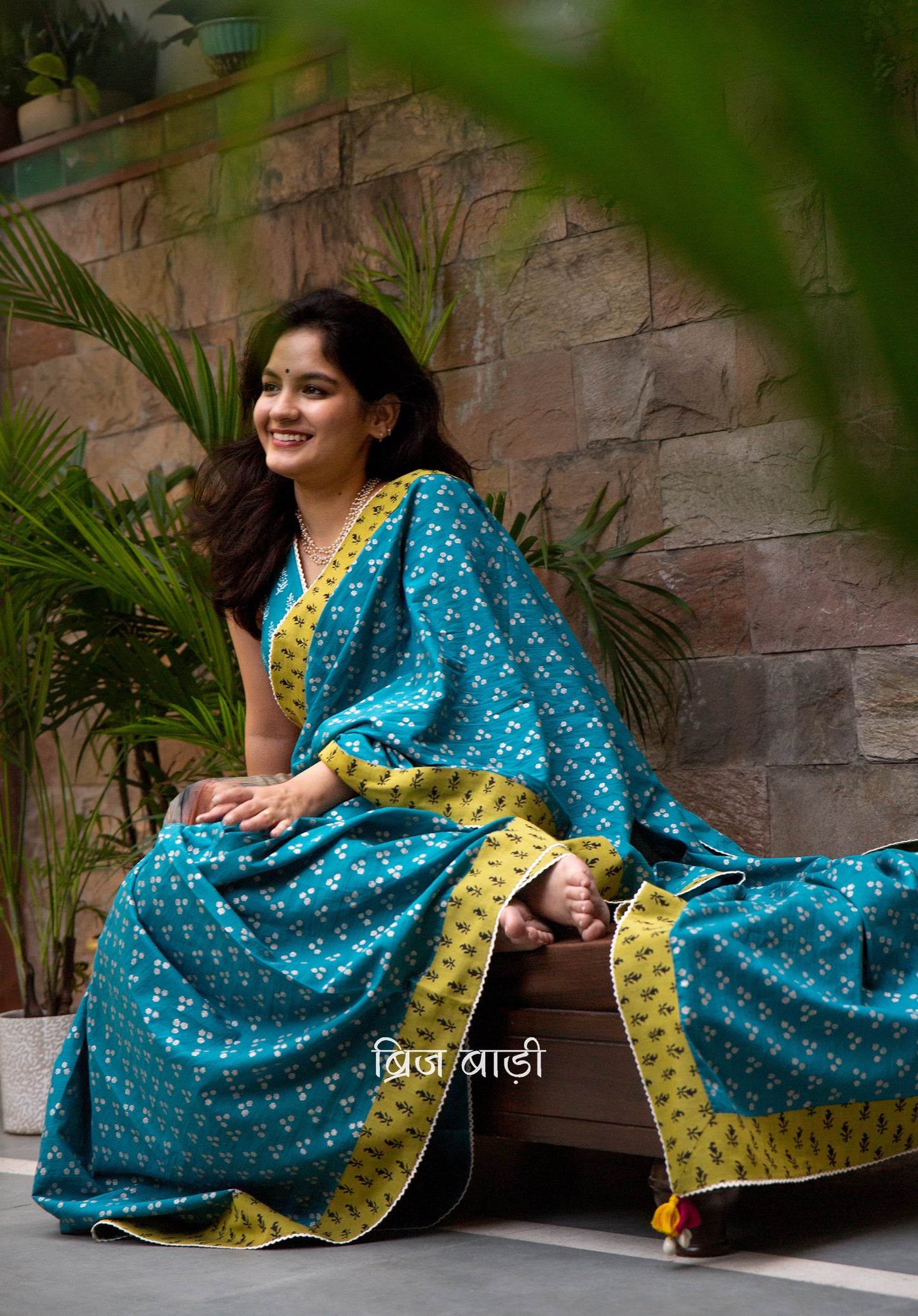 Teal blue cotton saree by Brij Bari, hand block printed with silver polka dot motifs & detailed with contrasting light green block printed border. Accented with delicate crochet lace that adds to its handcrafted charm. The pallu is finished with artisanal yellow & pink handmade thread tassels & tiny sea shells on the corner infusing it with a joyful festive flair. Comes with a free block printed blouse piece. A perfect blend of comfort & striking elegance for festive gatherings. Free blouse piece & shipping