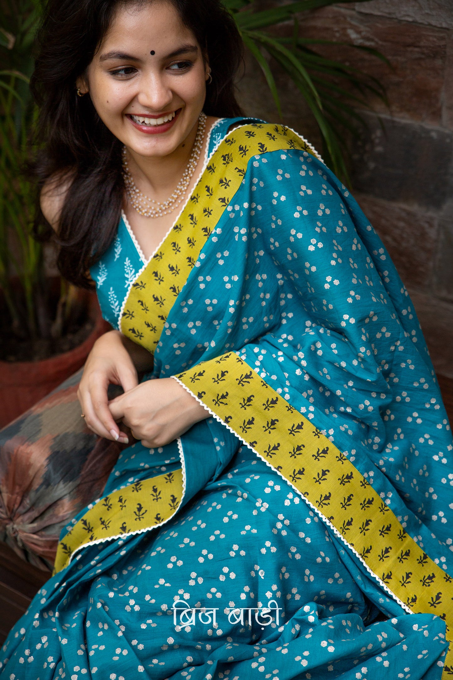 Teal blue cotton saree by Brij Bari, hand block printed with silver polka dot motifs & detailed with contrasting light green block printed border. Accented with delicate crochet lace that adds to its handcrafted charm. The pallu is finished with artisanal yellow & pink handmade thread tassels & tiny sea shells on the corner infusing it with a joyful festive flair. Comes with a free block printed blouse piece. A perfect blend of comfort & striking elegance for festive gatherings. Free blouse piece & shipping