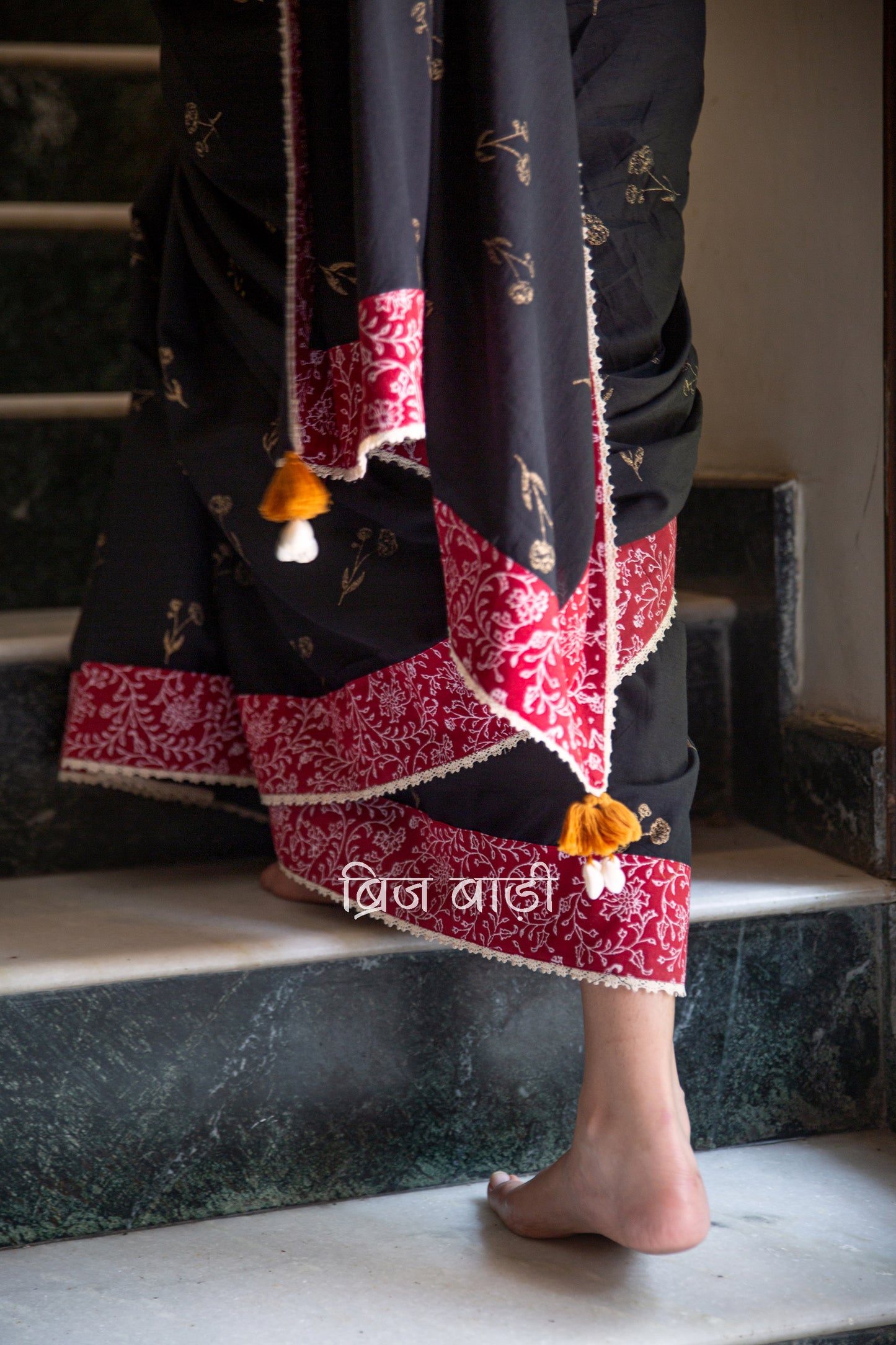 Black cotton saree by Brij Bari hand block printed with delicate gold floral motifs for an elegant festive look. The saree is enhanced with a contrasting deep red block printed border & finished with intricate crochet lace, blending traditional charm with handcrafted finesse. The pallu has  mustard handmade thread tassels, each accented with sea shells at the corner. Perfect for festive gatherings or special occasions, saree ensures you stand out & be comfortable. Free Shipping pan India. Free Blouse piece