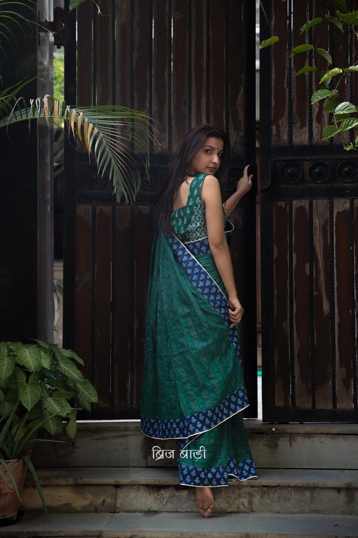Dark green cotton saree by Brij Bari, crafted for festive elegance and all-day comfort. Adorned with subtle lurex threads and enriched with a hand block printed blue border. Finished with artisanal crochet lace for a refined handmade touch. The pallu is delicately adorned with red handmade thread tassels and tiny sea shells at the corner, adding a soulful, crafted character. A perfect pick for festive gatherings and special occasions. Includes a free block printed blouse piece. Free shipping pan India.