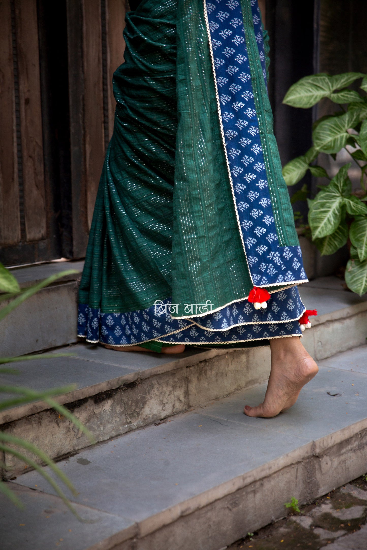Dark green cotton saree by Brij Bari, crafted for festive elegance and all-day comfort. Adorned with subtle lurex threads and enriched with a hand block printed blue border. Finished with artisanal crochet lace for a refined handmade touch. The pallu is delicately adorned with red handmade thread tassels and tiny sea shells at the corner, adding a soulful, crafted character. A perfect pick for festive gatherings and special occasions. Includes a free block printed blouse piece. Free shipping pan India.
