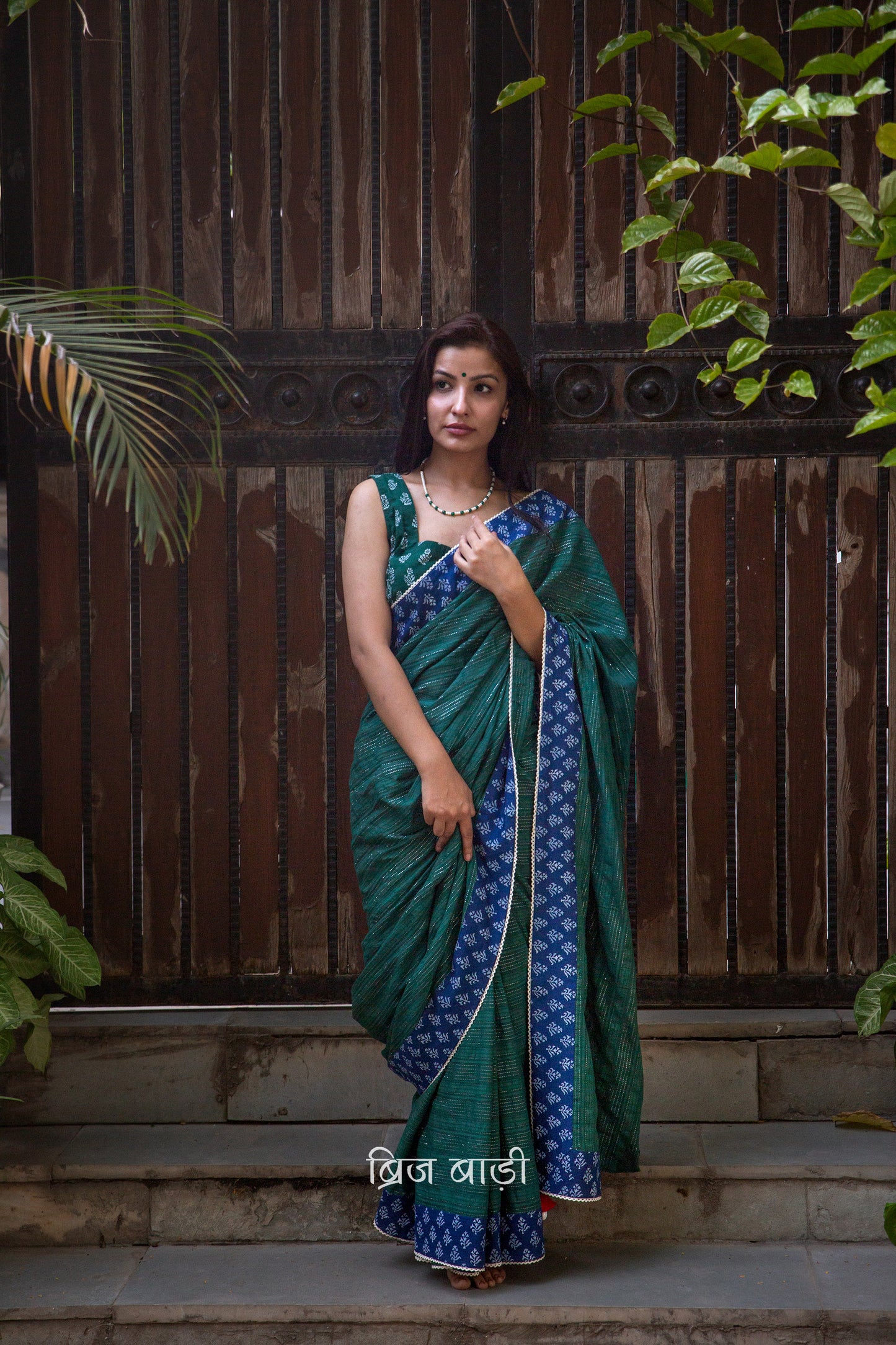 Dark green cotton saree by Brij Bari, crafted for festive elegance and all-day comfort. Adorned with subtle lurex threads and enriched with a hand block printed blue border. Finished with artisanal crochet lace for a refined handmade touch. The pallu is delicately adorned with red handmade thread tassels and tiny sea shells at the corner, adding a soulful, crafted character. A perfect pick for festive gatherings and special occasions. Includes a free block printed blouse piece. Free shipping pan India.