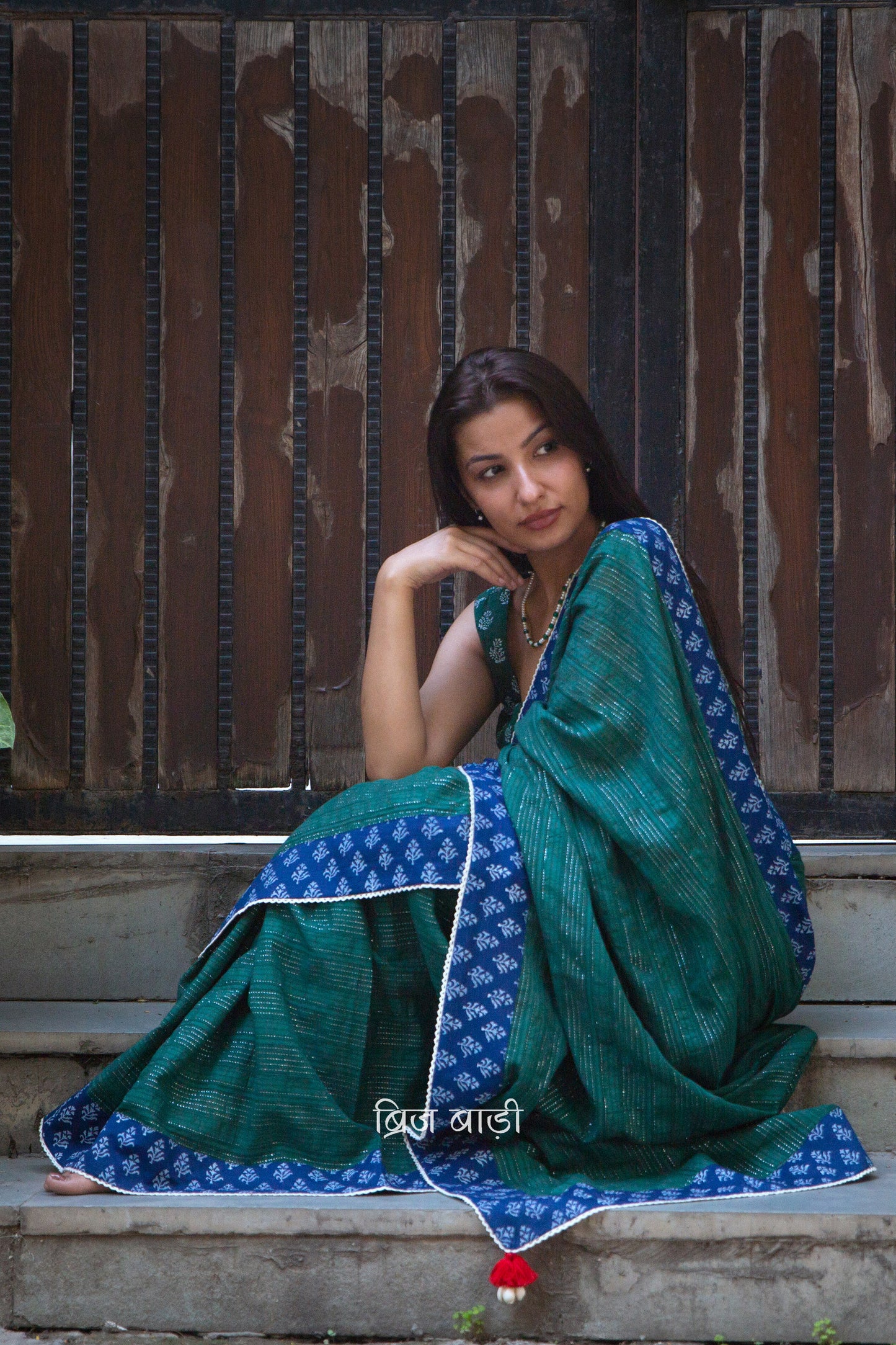Dark green cotton saree by Brij Bari, crafted for festive elegance and all-day comfort. Adorned with subtle lurex threads and enriched with a hand block printed blue border. Finished with artisanal crochet lace for a refined handmade touch. The pallu is delicately adorned with red handmade thread tassels and tiny sea shells at the corner, adding a soulful, crafted character. A perfect pick for festive gatherings and special occasions. Includes a free block printed blouse piece. Free shipping pan India.