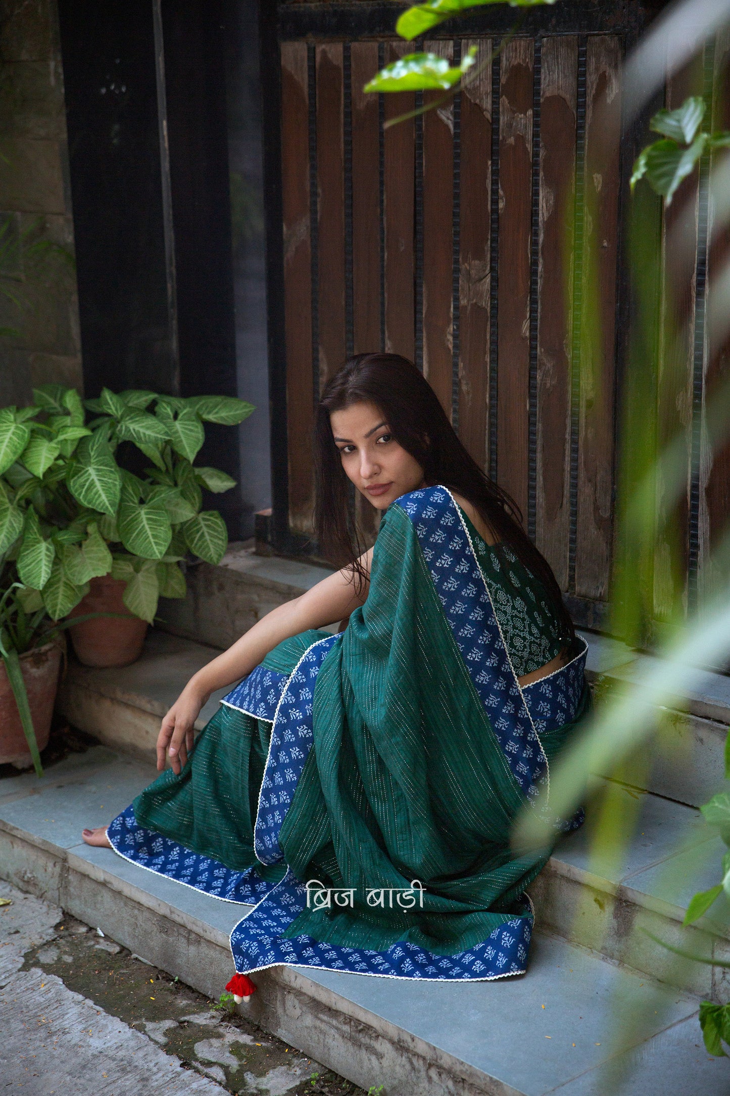 Dark green cotton saree by Brij Bari, crafted for festive elegance and all-day comfort. Adorned with subtle lurex threads and enriched with a hand block printed blue border. Finished with artisanal crochet lace for a refined handmade touch. The pallu is delicately adorned with red handmade thread tassels and tiny sea shells at the corner, adding a soulful, crafted character. A perfect pick for festive gatherings and special occasions. Includes a free block printed blouse piece. Free shipping pan India.