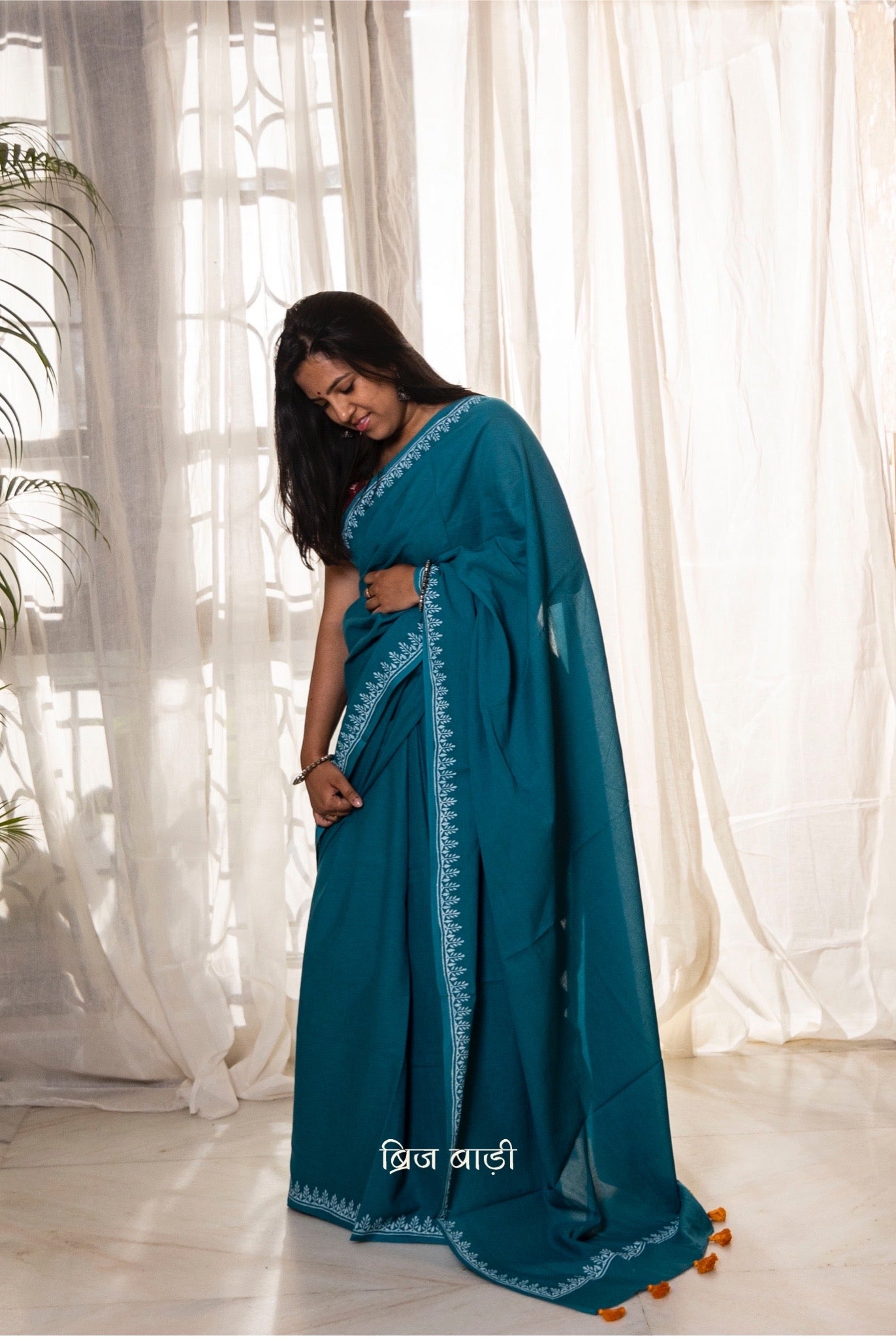Teal blue soft and light-weight cotton saree by Brij Bari, designed for all-day comfort and effortless elegance. Detailed with a delicate hand block printed border in crisp white, creating a refined contrast. The saree is finished with orange handmade thread tassels on the pallu, adding a subtle pop of colour. Comes with a free matching block printed blouse piece. Ideal for everyday wear, office, casual outings, or light festive occasions, with free shipping pan India.