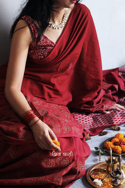 Deep red light-weight cotton saree by Brij Bari, featuring intricate rose block printed motifs in golden on the skirt and elegant geometric patterns on the pallu in silver, lending a festive and graceful touch. The pallu is finished with handcrafted olive green thread tassels that add earthy contrast and texture. Includes a free hand block printed blouse piece in matching tones. A perfect blend of festive elegance and artisan detail, crafted with care by Brij Bari.