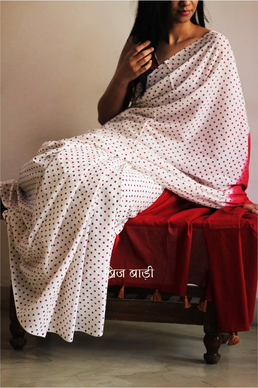 Off-white soft and light-weight cotton saree by Brij Bari, featuring deep red ombre pallu and small retro-style polka dot hand block prints. Accented with handcrafted mustard thread tassels on the pallu for a touch of artisanal charm. Includes a free matching hand block printed blouse piece. A timeless piece combining elegance, tradition, and comfort—crafted by Brij Bari’s skilled artisans.