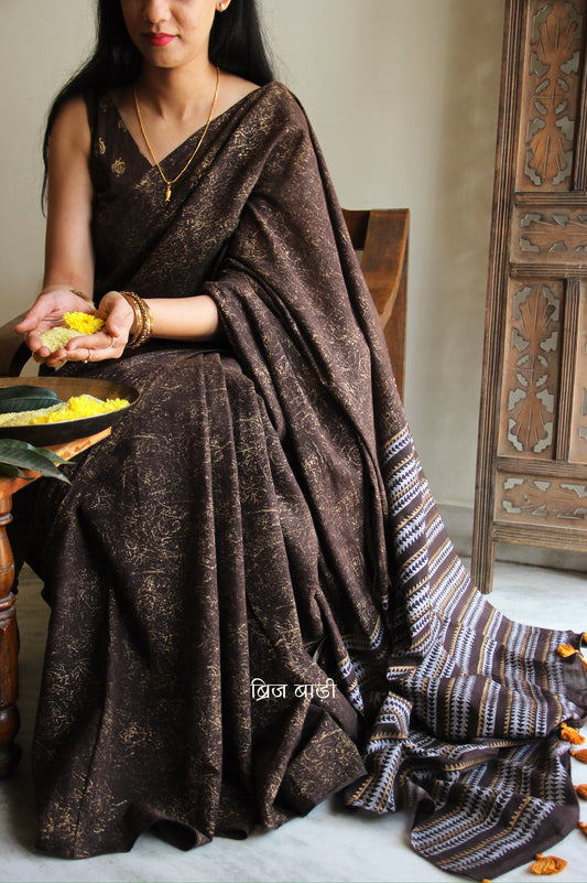 Brown soft cotton saree by Brij Bari featuring a striking hand block printed abstract pattern in gold across the body, paired with a contemporary geometric design in silver and gold on the pallu. This blend of traditional technique and modern aesthetics gives the saree a unique festive appeal. The pallu is finished with handcrafted mustard thread tassels that add a warm, artisanal touch. Includes a free matching hand block printed blouse piece. A graceful and comfortable choice for festive gatherings