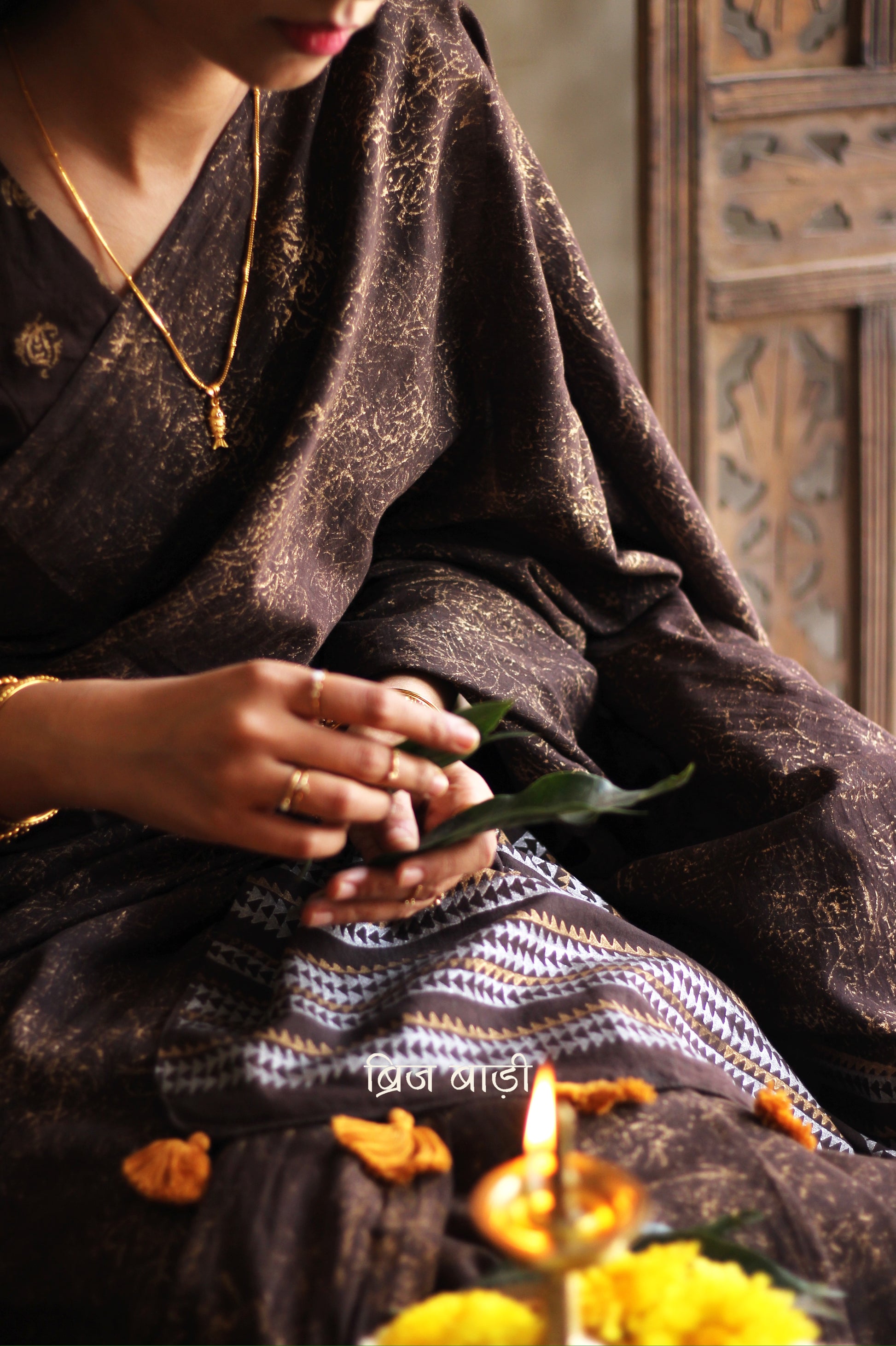 Brown soft cotton saree by Brij Bari featuring a striking hand block printed abstract pattern in gold across the body, paired with a contemporary geometric design in silver and gold on the pallu. This blend of traditional technique and modern aesthetics gives the saree a unique festive appeal. The pallu is finished with handcrafted mustard thread tassels that add a warm, artisanal touch. Includes a free matching hand block printed blouse piece. A graceful and comfortable choice for festive gatherings