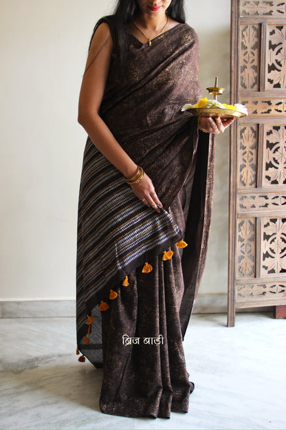 Brown soft cotton saree by Brij Bari featuring a striking hand block printed abstract pattern in gold across the body, paired with a contemporary geometric design in silver and gold on the pallu. This blend of traditional technique and modern aesthetics gives the saree a unique festive appeal. The pallu is finished with handcrafted mustard thread tassels that add a warm, artisanal touch. Includes a free matching hand block printed blouse piece. A graceful and comfortable choice for festive gatherings
