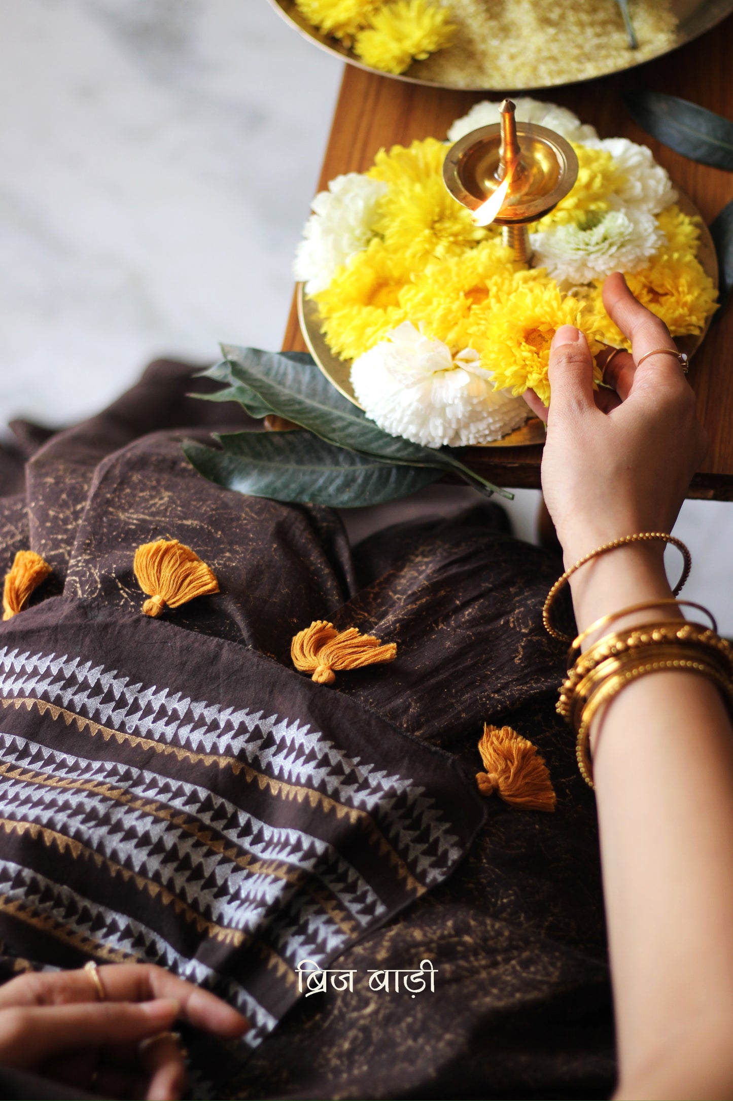 Brown soft cotton saree by Brij Bari featuring a striking hand block printed abstract pattern in gold across the body, paired with a contemporary geometric design in silver and gold on the pallu. This blend of traditional technique and modern aesthetics gives the saree a unique festive appeal. The pallu is finished with handcrafted mustard thread tassels that add a warm, artisanal touch. Includes a free matching hand block printed blouse piece. A graceful and comfortable choice for festive gatherings