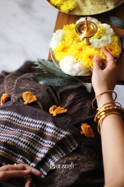 Brown soft cotton saree by Brij Bari featuring a striking hand block printed abstract pattern in gold across the body, paired with a contemporary geometric design in silver and gold on the pallu. This blend of traditional technique and modern aesthetics gives the saree a unique festive appeal. The pallu is finished with handcrafted mustard thread tassels that add a warm, artisanal touch. Includes a free matching hand block printed blouse piece. A graceful and comfortable choice for festive gatherings