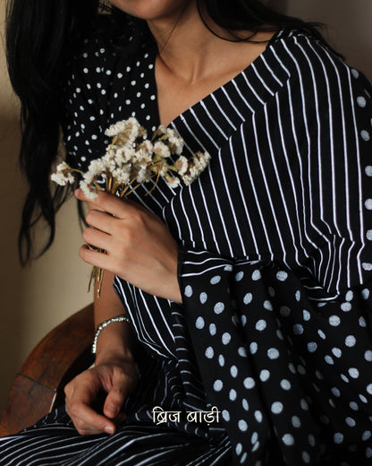 Black soft cotton saree by Brij Bari, featuring a bold and striking hand block printed pattern of stripes and polka dots for a modern, playful twist. Crafted from breathable, light-weight cotton, the saree drapes effortlessly—ideal for both everyday wear and special occasions. Finished with handcrafted red thread tassels on the pallu, adding an artisanal touch of charm. It comes with a free hand block printed blouse piece that complements the print and palette. 
