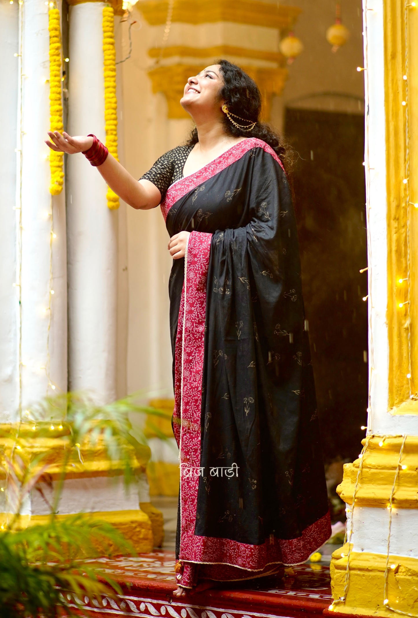 Black cotton saree by Brij Bari hand block printed with delicate gold floral motifs for an elegant festive look. The saree is enhanced with a contrasting deep red block printed border & finished with intricate crochet lace, blending traditional charm with handcrafted finesse. The pallu has  mustard handmade thread tassels, each accented with sea shells at the corner. Perfect for festive gatherings or special occasions, saree ensures you stand out & be comfortable. Free Shipping pan India. Free Blouse piece