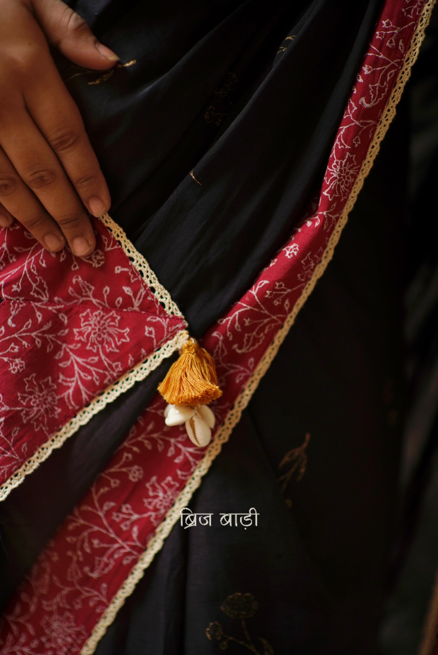 Black cotton saree by Brij Bari hand block printed with delicate gold floral motifs for an elegant festive look. The saree is enhanced with a contrasting deep red block printed border & finished with intricate crochet lace, blending traditional charm with handcrafted finesse. The pallu has  mustard handmade thread tassels, each accented with sea shells at the corner. Perfect for festive gatherings or special occasions, saree ensures you stand out & be comfortable. Free Shipping pan India. Free Blouse piece