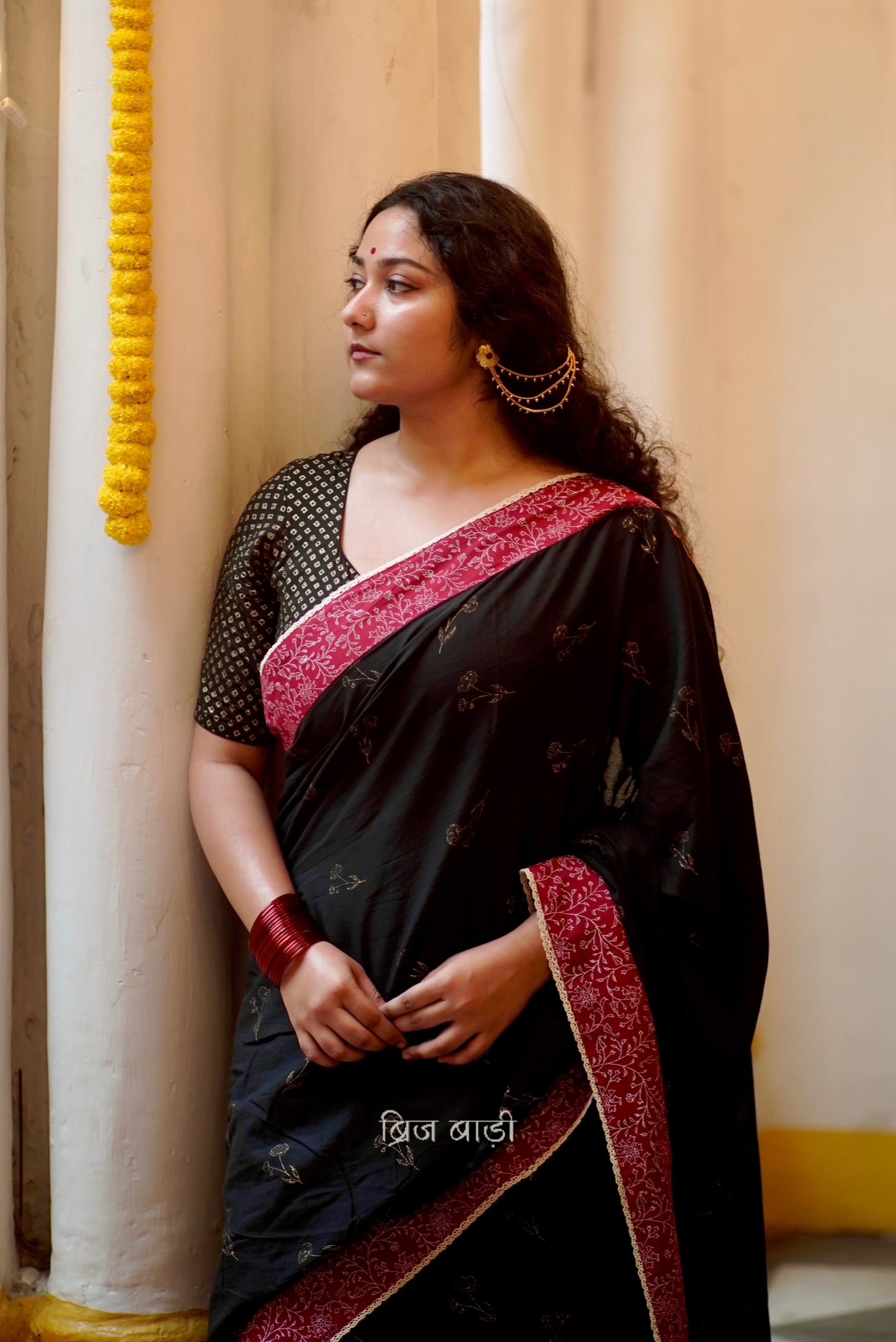 Black cotton saree by Brij Bari hand block printed with delicate gold floral motifs for an elegant festive look. The saree is enhanced with a contrasting deep red block printed border & finished with intricate crochet lace, blending traditional charm with handcrafted finesse. The pallu has  mustard handmade thread tassels, each accented with sea shells at the corner. Perfect for festive gatherings or special occasions, saree ensures you stand out & be comfortable. Free Shipping pan India. Free Blouse piece
