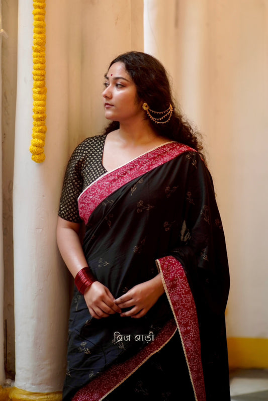 Black cotton saree by Brij Bari hand block printed with delicate gold floral motifs for an elegant festive look. The saree is enhanced with a contrasting deep red block printed border & finished with intricate crochet lace, blending traditional charm with handcrafted finesse. The pallu has  mustard handmade thread tassels, each accented with sea shells at the corner. Perfect for festive gatherings or special occasions, saree ensures you stand out & be comfortable. Free Shipping pan India. Free Blouse piece