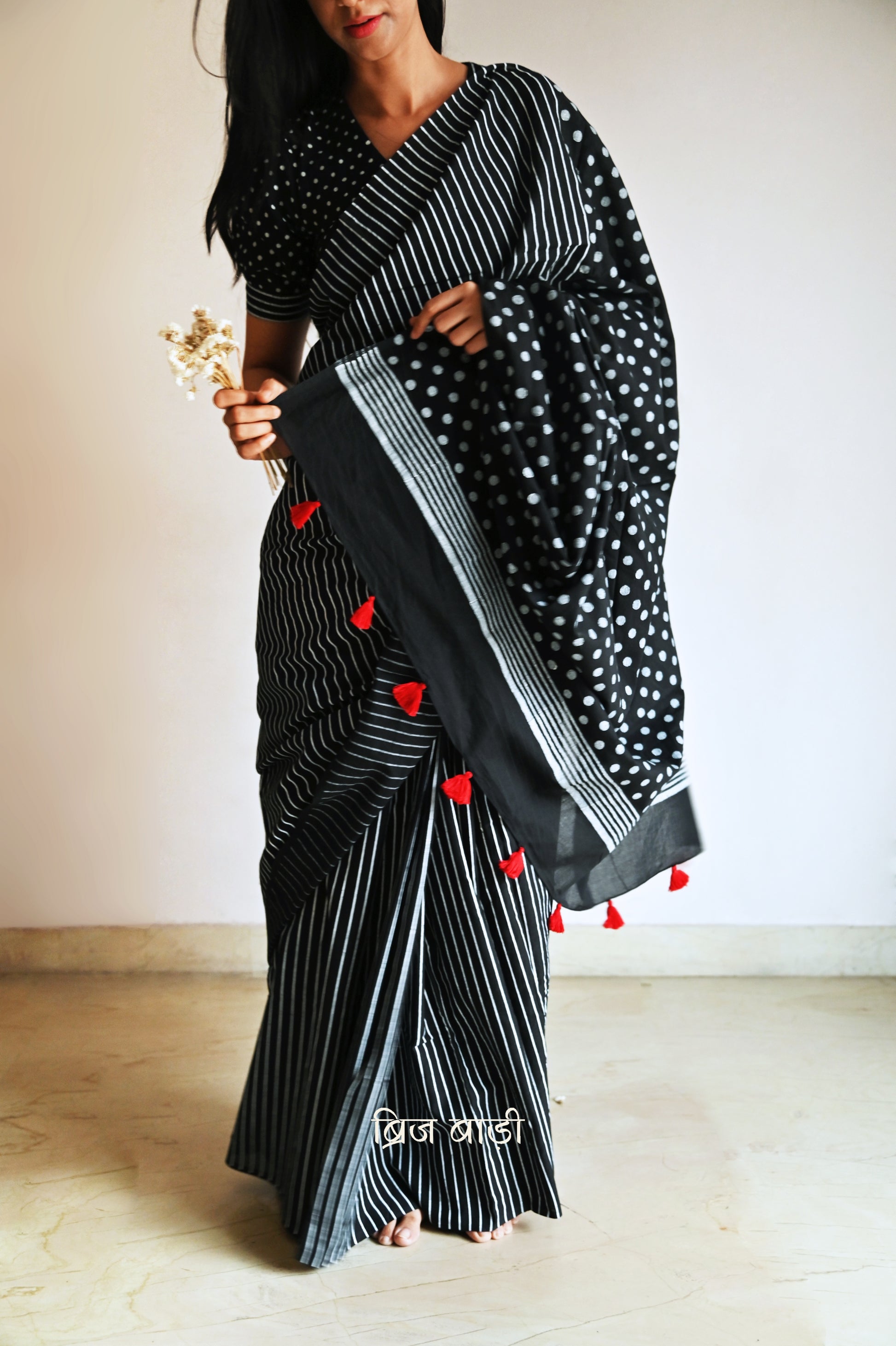 Black soft cotton saree by Brij Bari, featuring a bold and striking hand block printed pattern of stripes and polka dots for a modern, playful twist. Handcrafted from breathable, light-weight cotton, the saree drapes effortlessly—ideal for both everyday wear and special occasions. Finished with handcrafted red thread tassels on the pallu, adding an artisanal touch of charm. It comes with a free hand block printed blouse piece that complements the print and palette. 